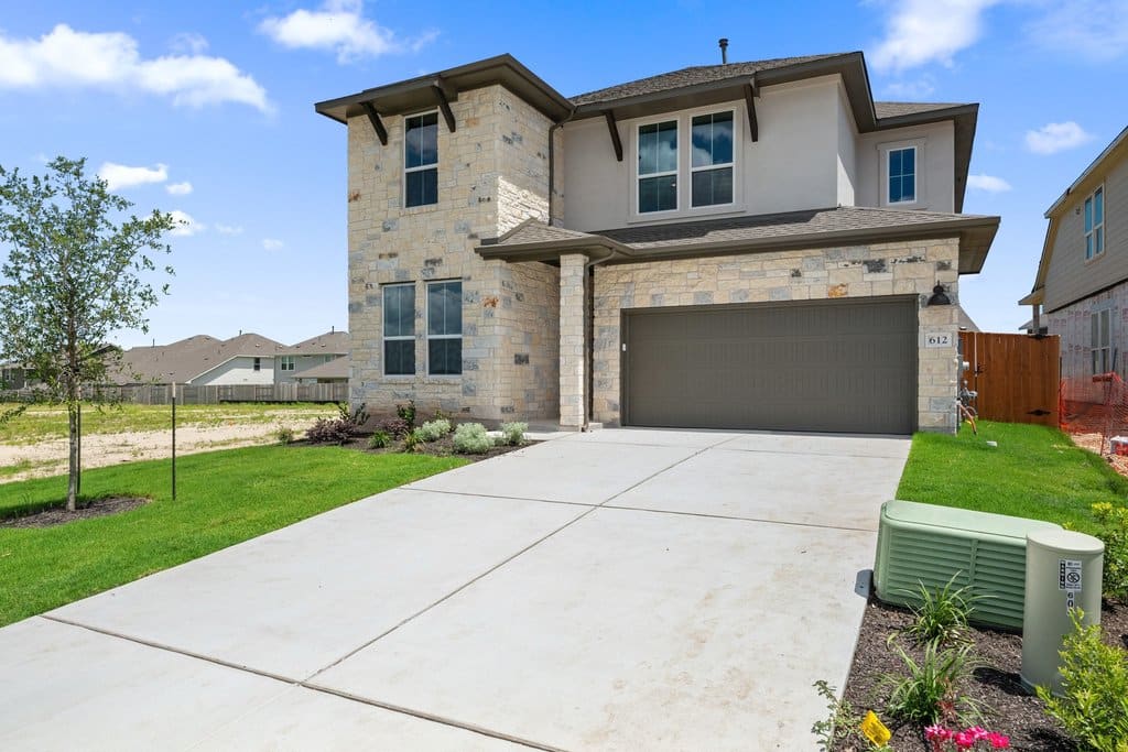 A modern two-story house with a stone facade and a gray garage door sits on a concrete driveway, with a neatly mowed lawn and a young tree in the foreground, while other houses and a bright blue sky with scattered clouds form the background.