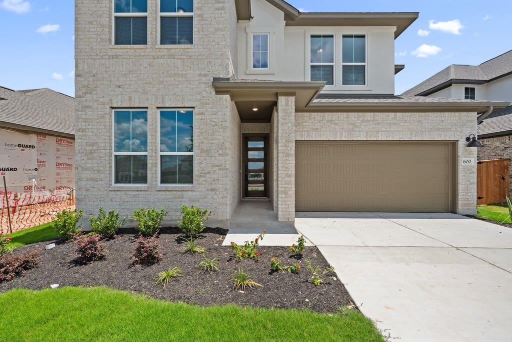 A modern two-story house with a light brick facade and a beige garage door is fronted by a landscaped garden bed, with other similar houses visible in the background.