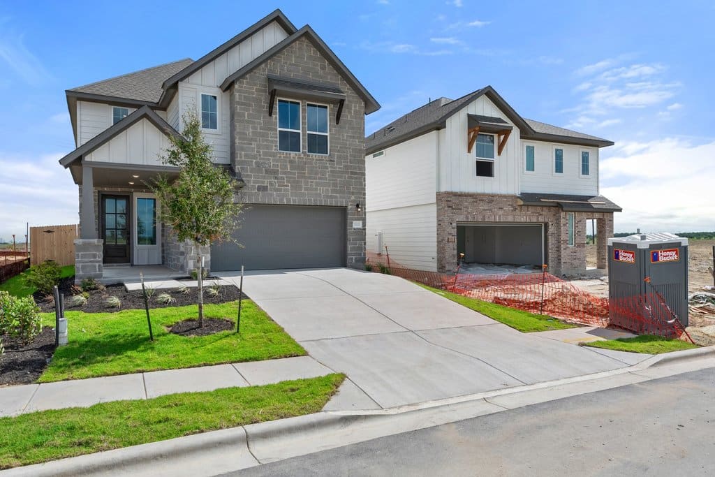 A well-maintained front yard with a paved driveway leads to two modern houses, one completed and the other under construction, with a bright blue sky overhead.
