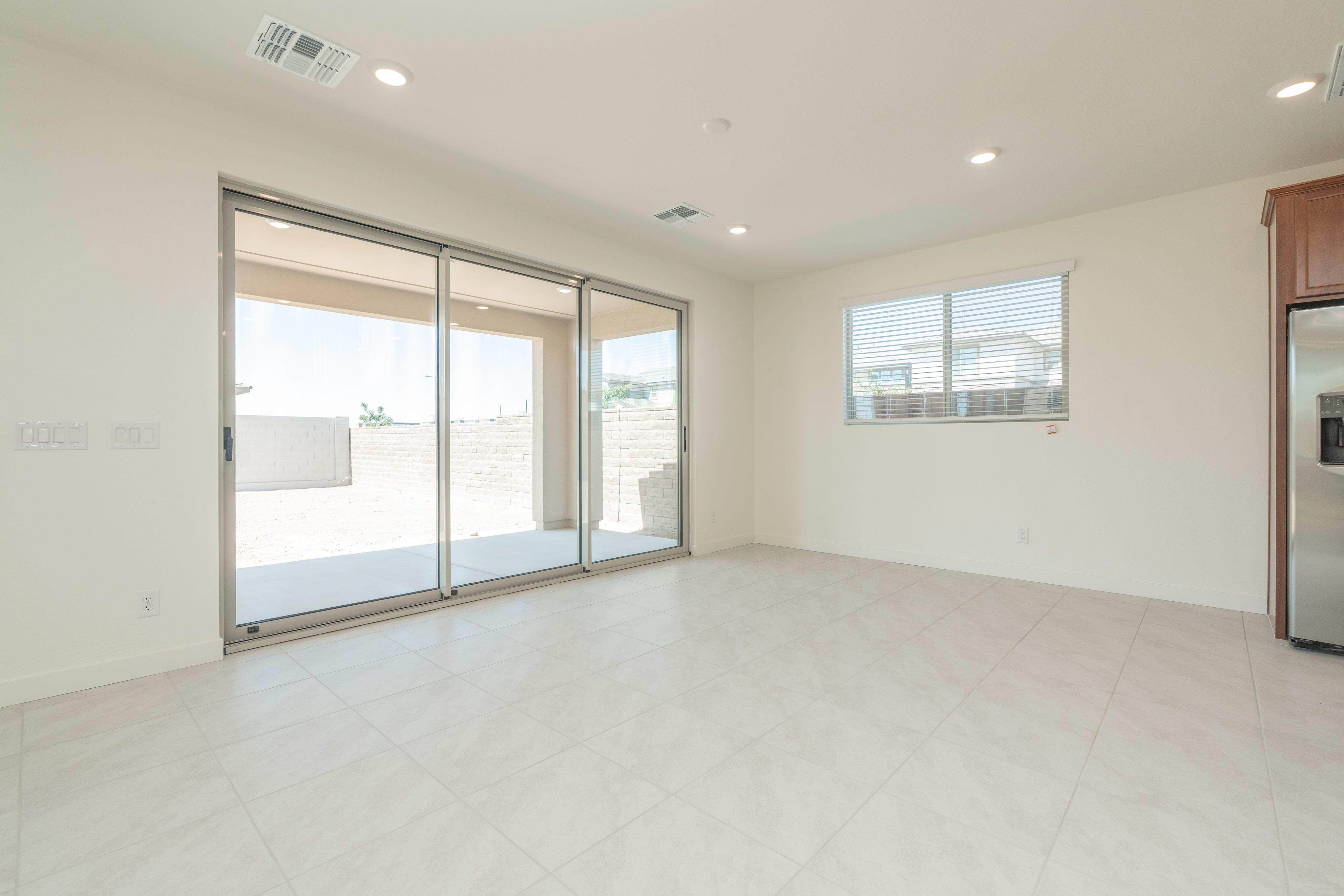 A bright, empty room with large sliding glass doors opening to a patio in the foreground, and a window overlooking houses in the background.