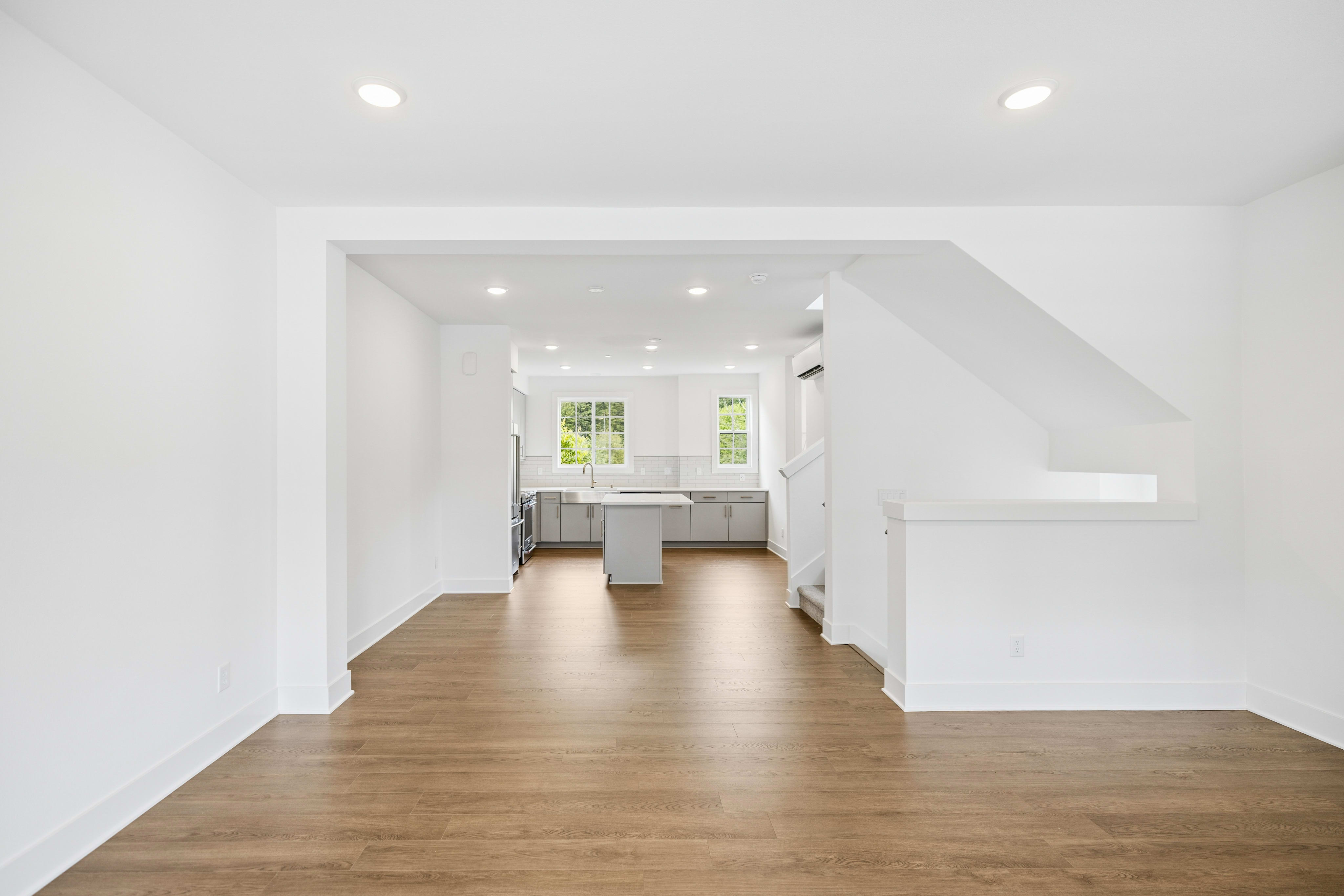 A bright, modern living room with hardwood floors opens to a spacious kitchen with gray cabinets and a central island, while a staircase ascends to the right.