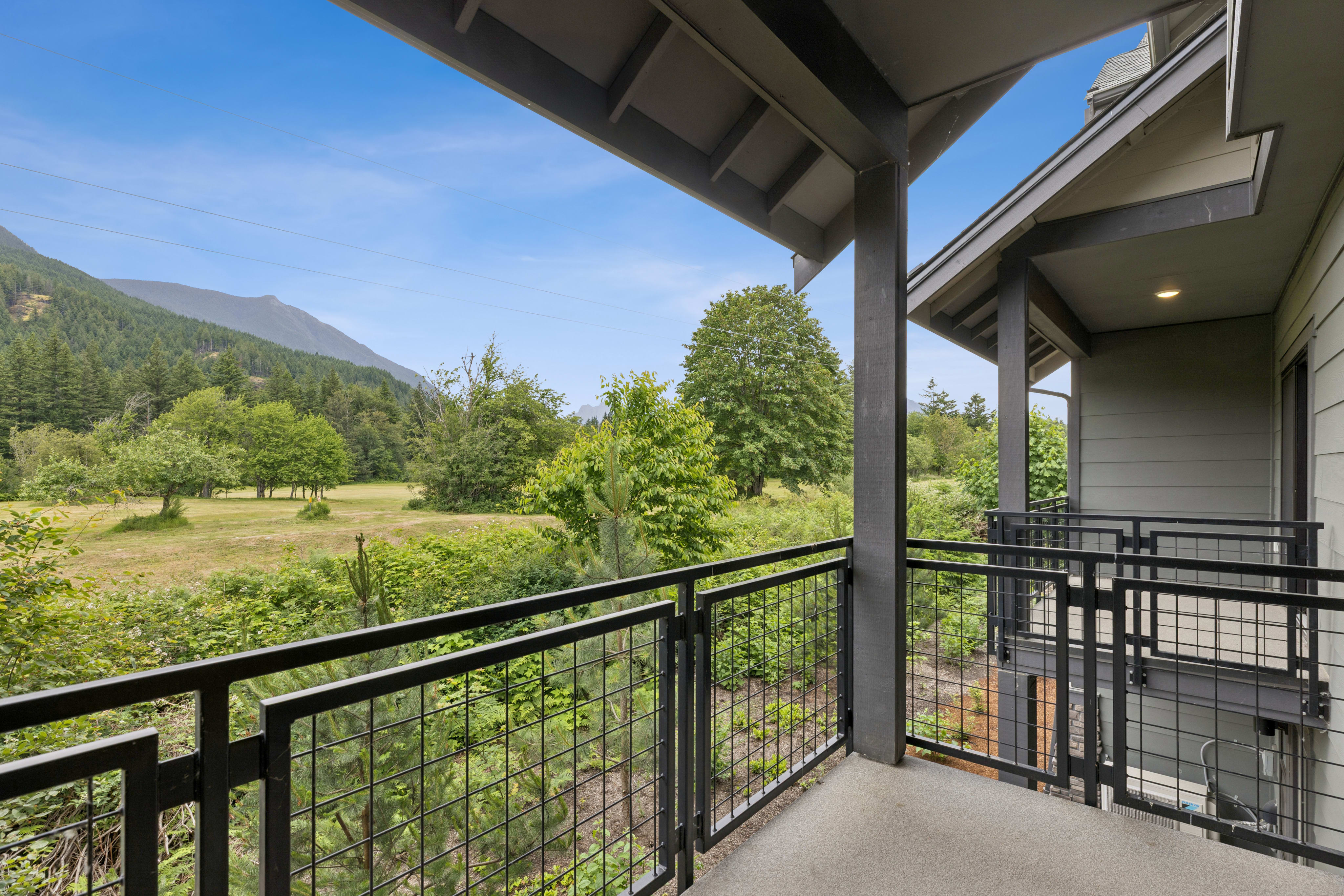 A modern balcony with a metal railing overlooks a lush green landscape with rolling hills and trees under a clear blue sky.