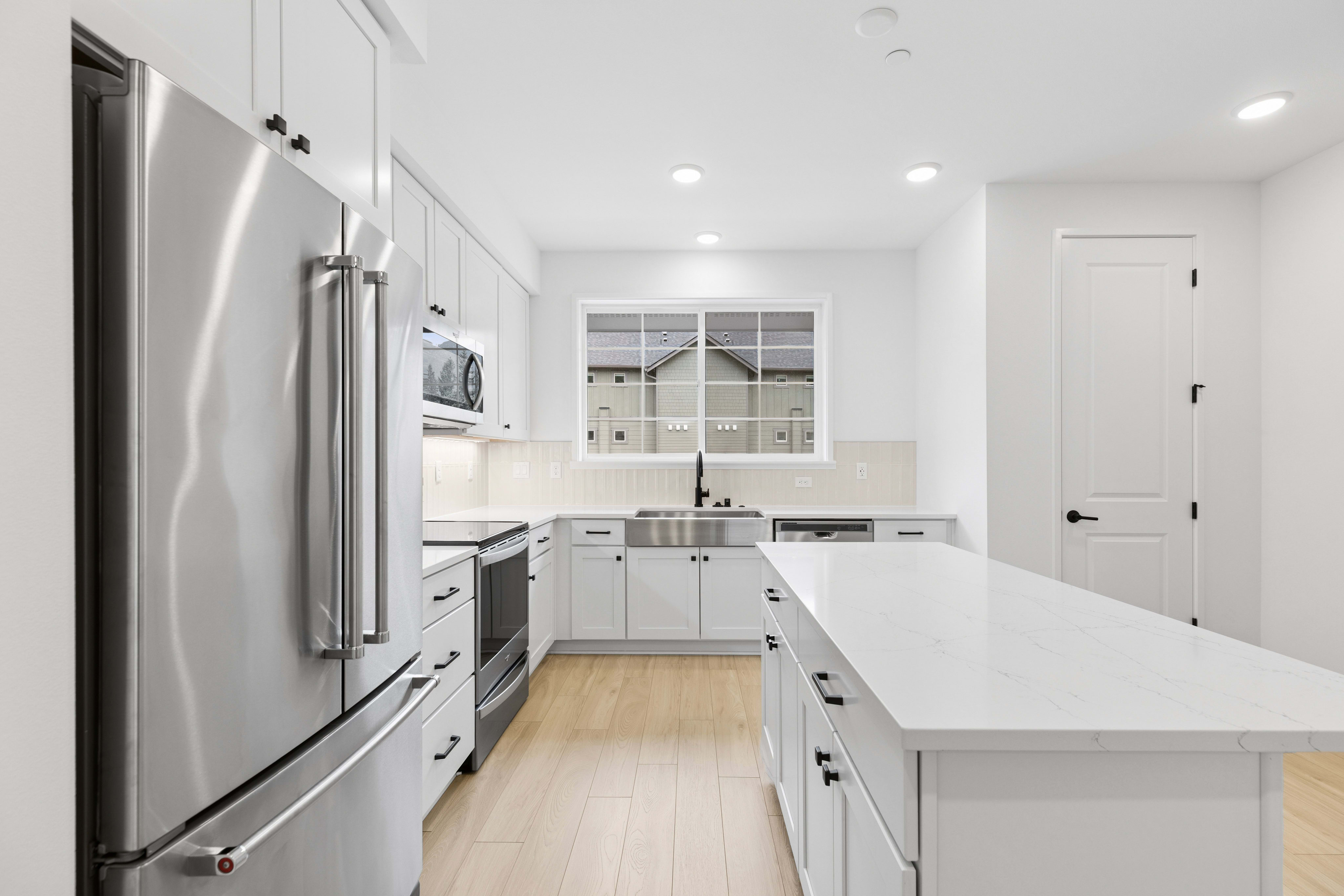 A modern white kitchen with a stainless steel refrigerator in the foreground and a large island, while the background features a sink under a window overlooking houses.