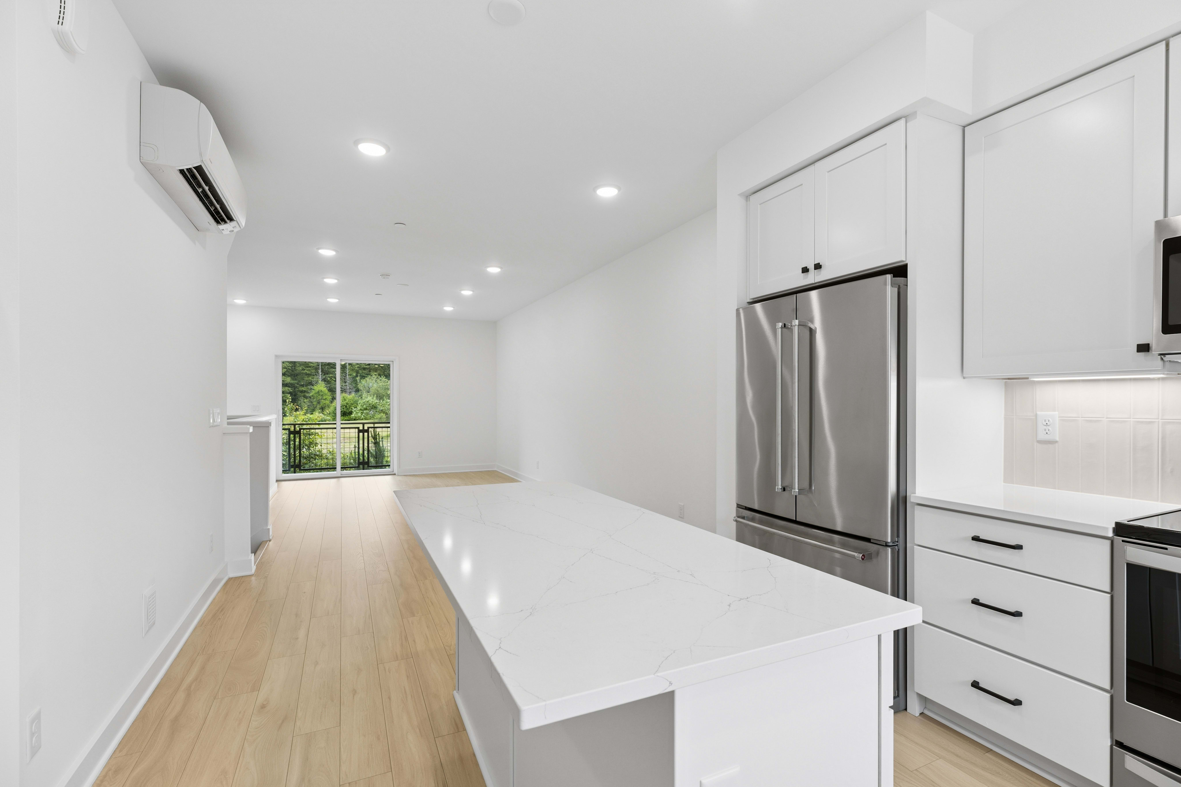 A modern white kitchen island with a stainless steel refrigerator in the foreground leads to a bright living area with large windows overlooking a green landscape in the background.