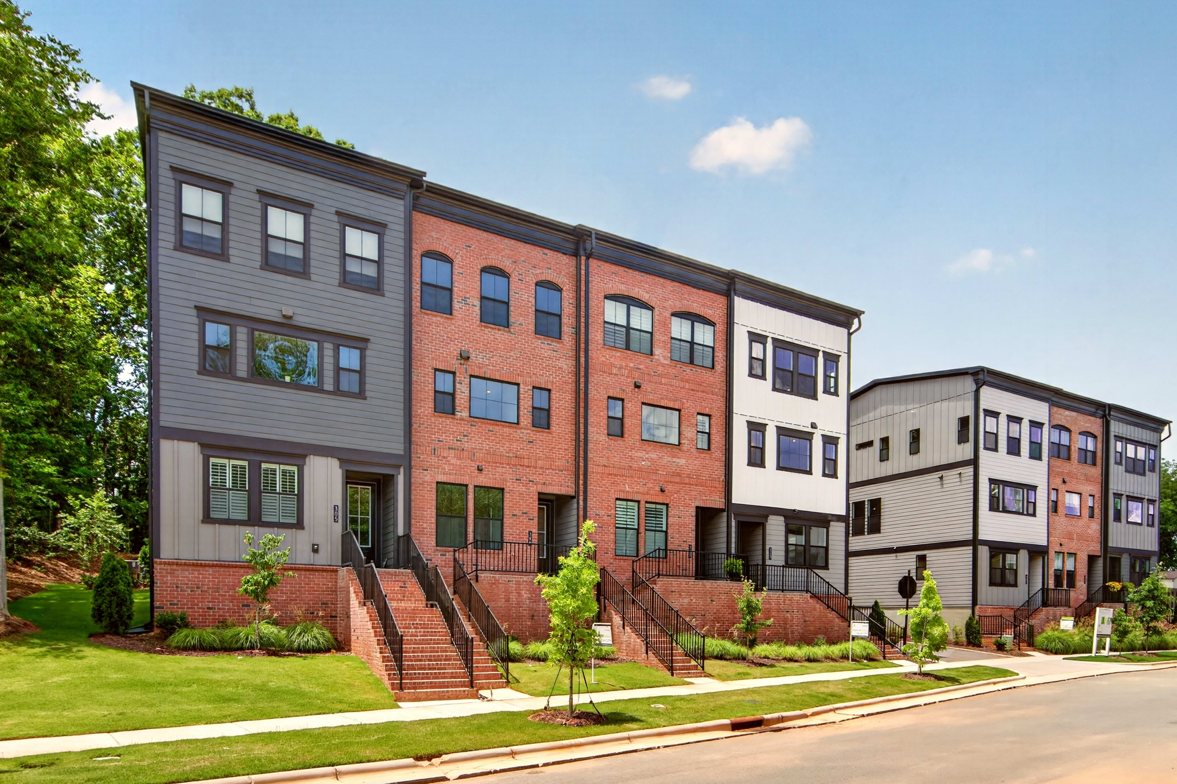 Modern townhouses with brick and gray siding stand on a grassy lawn in the foreground, with trees and a blue sky in the background.