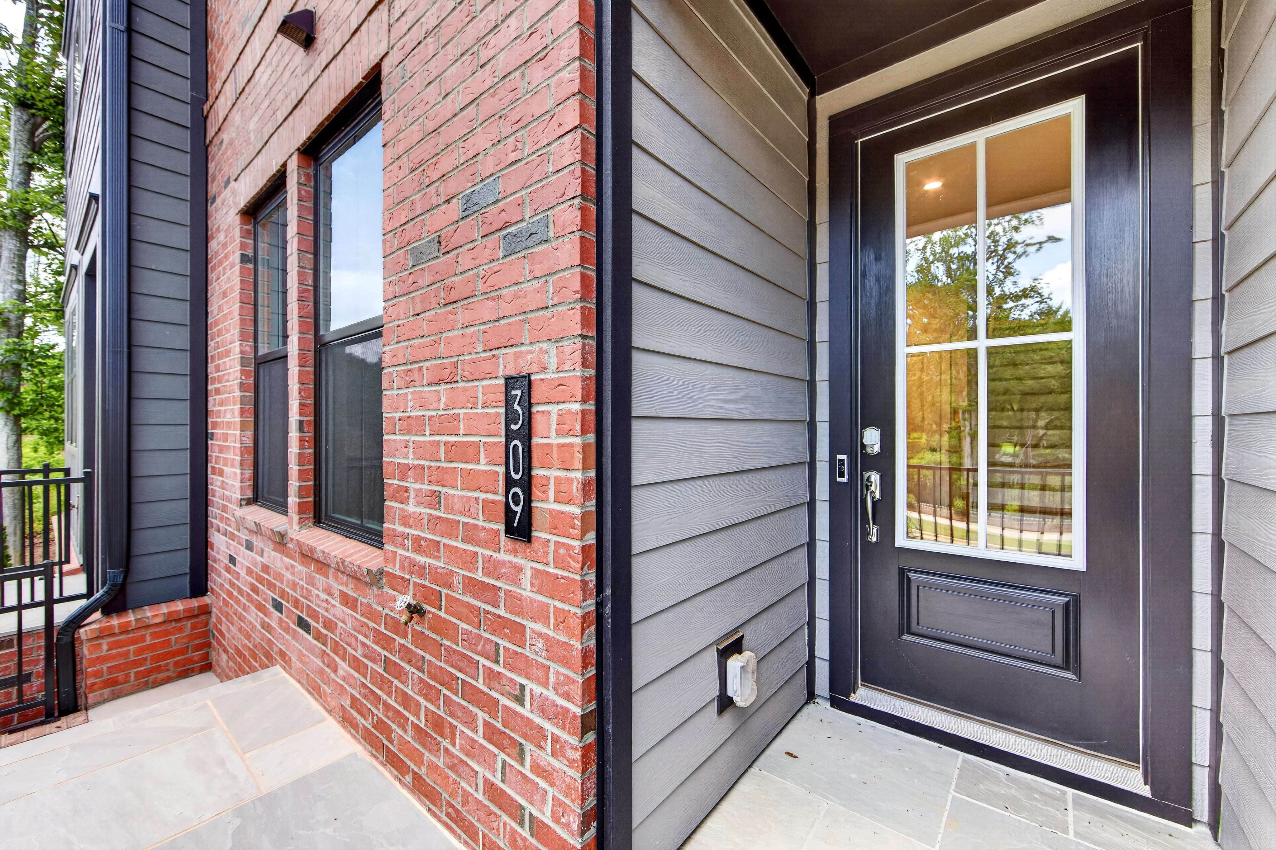 A modern townhouse entrance with a brick facade and gray siding is visible in the foreground, while lush green trees form the background.