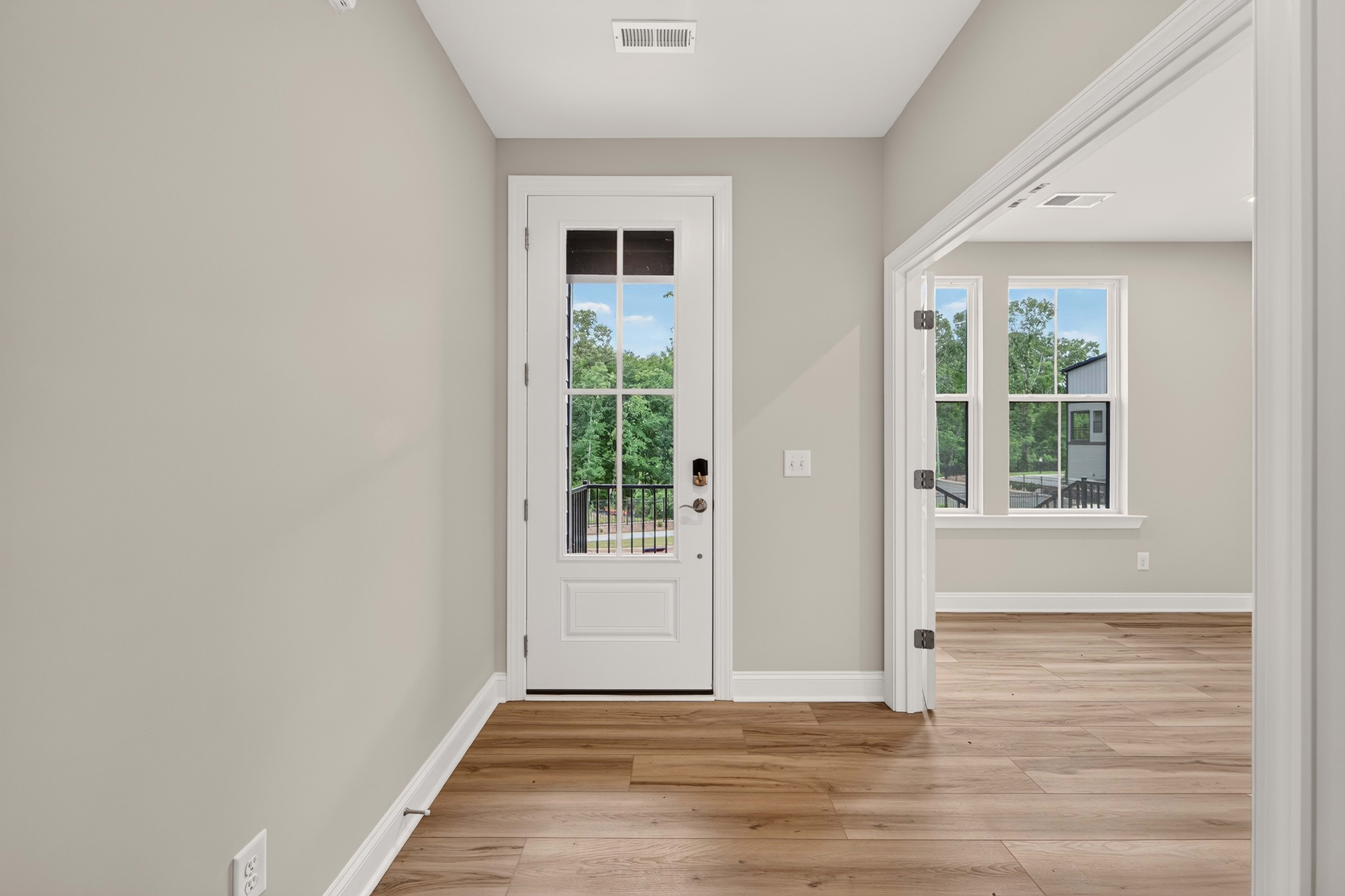 A white door with glass panes opens to a green, tree-filled outdoor space, while the foreground features a light-colored wall and wooden flooring.
