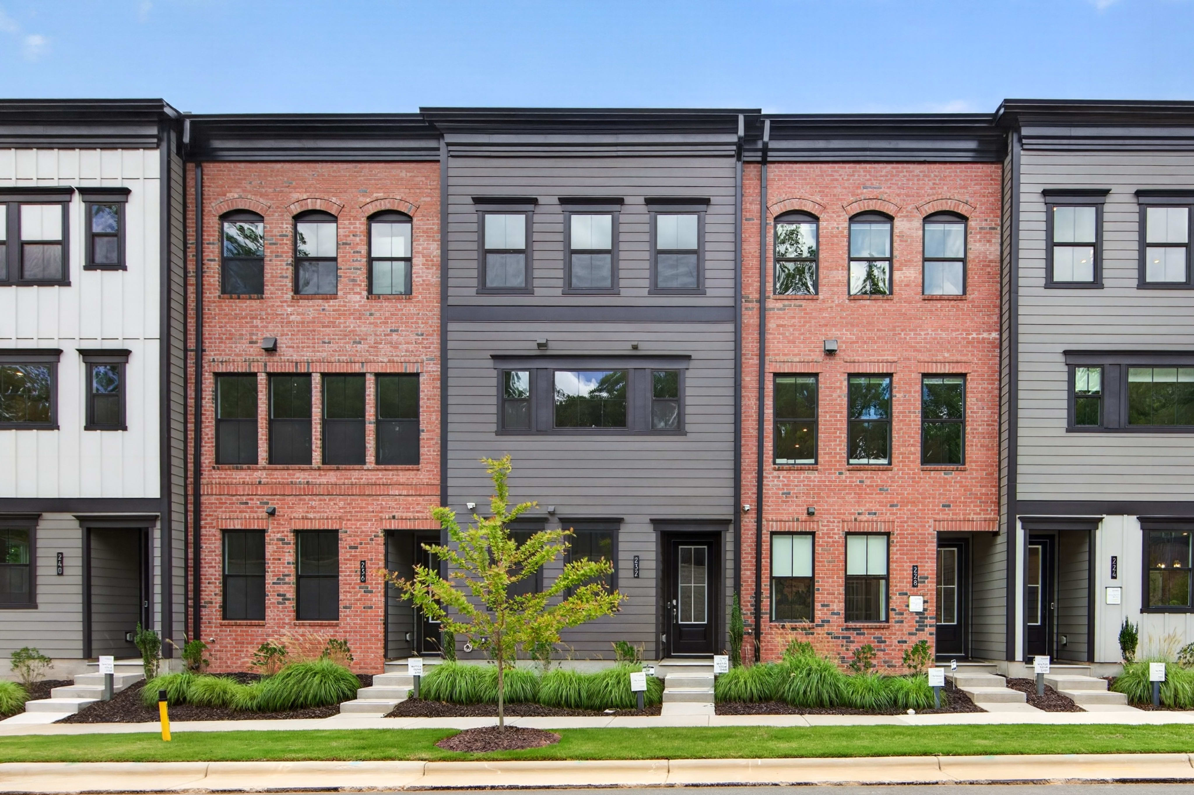 A row of modern townhouses with brick and gray siding stands behind a neatly landscaped lawn and a small tree.