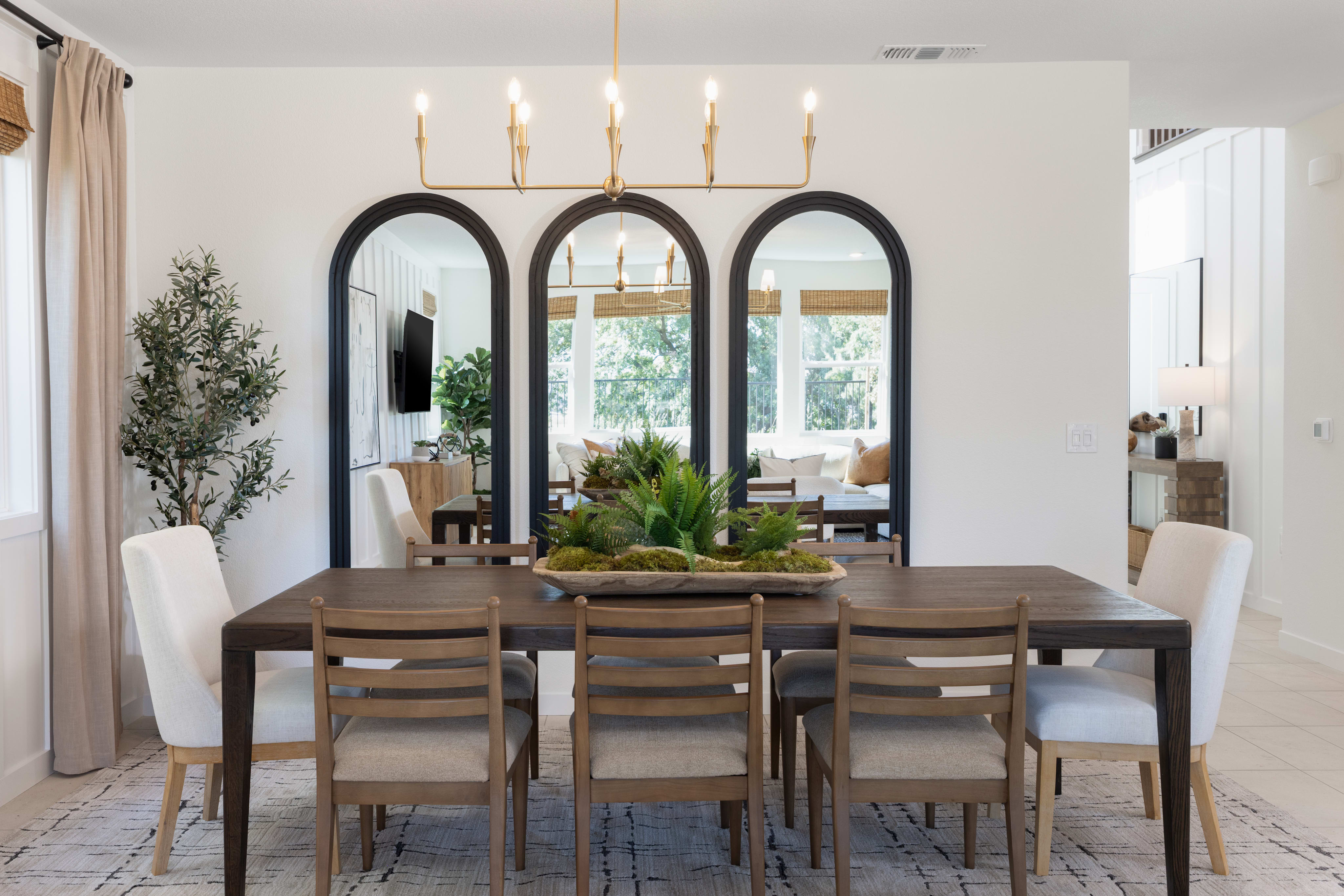 A dark wood dining table with chairs sits in the foreground, reflecting in three arched mirrors on a white wall in the background.