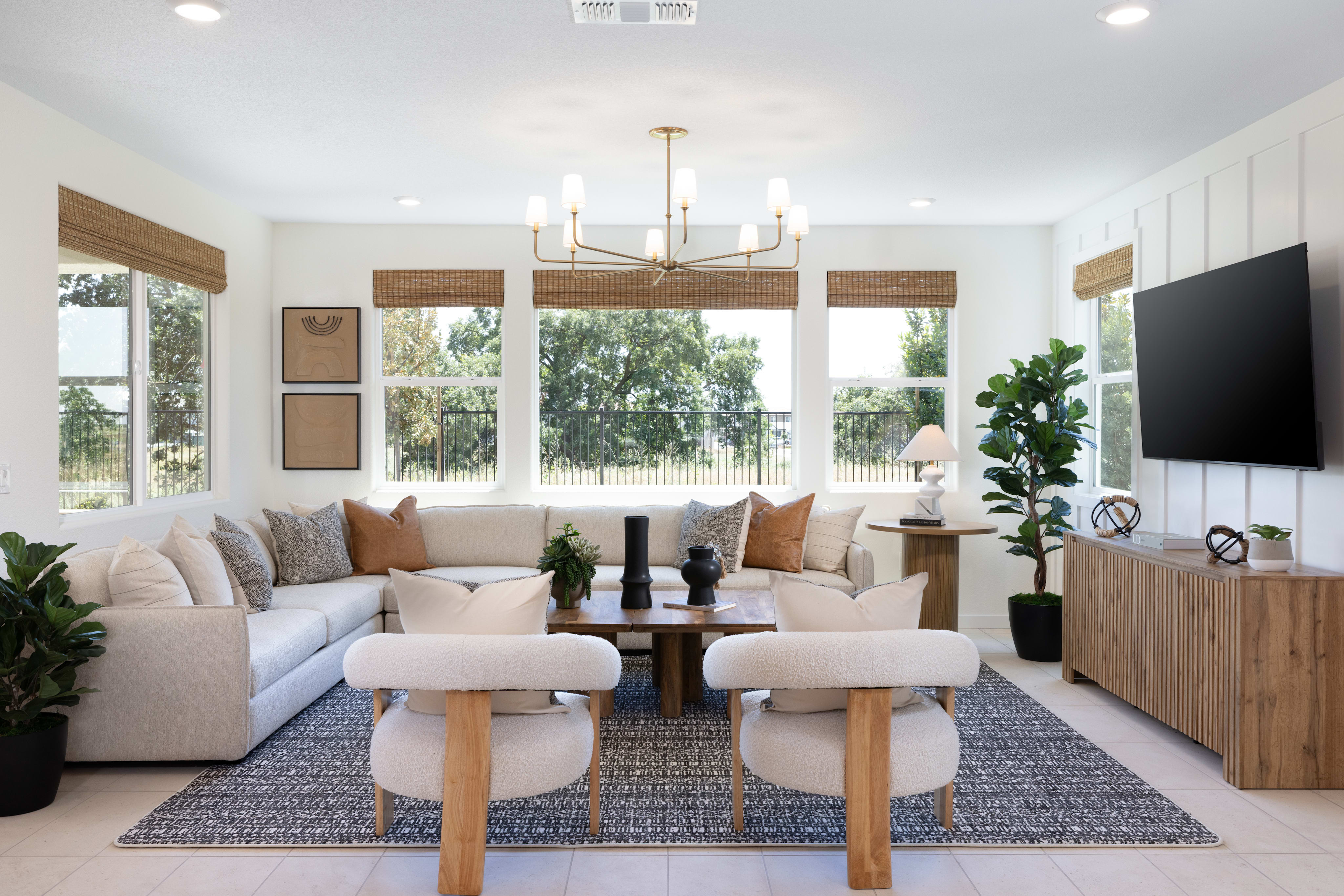 A modern living room with a sectional sofa and two armchairs in the foreground, and large windows overlooking a green, tree-filled landscape in the background.