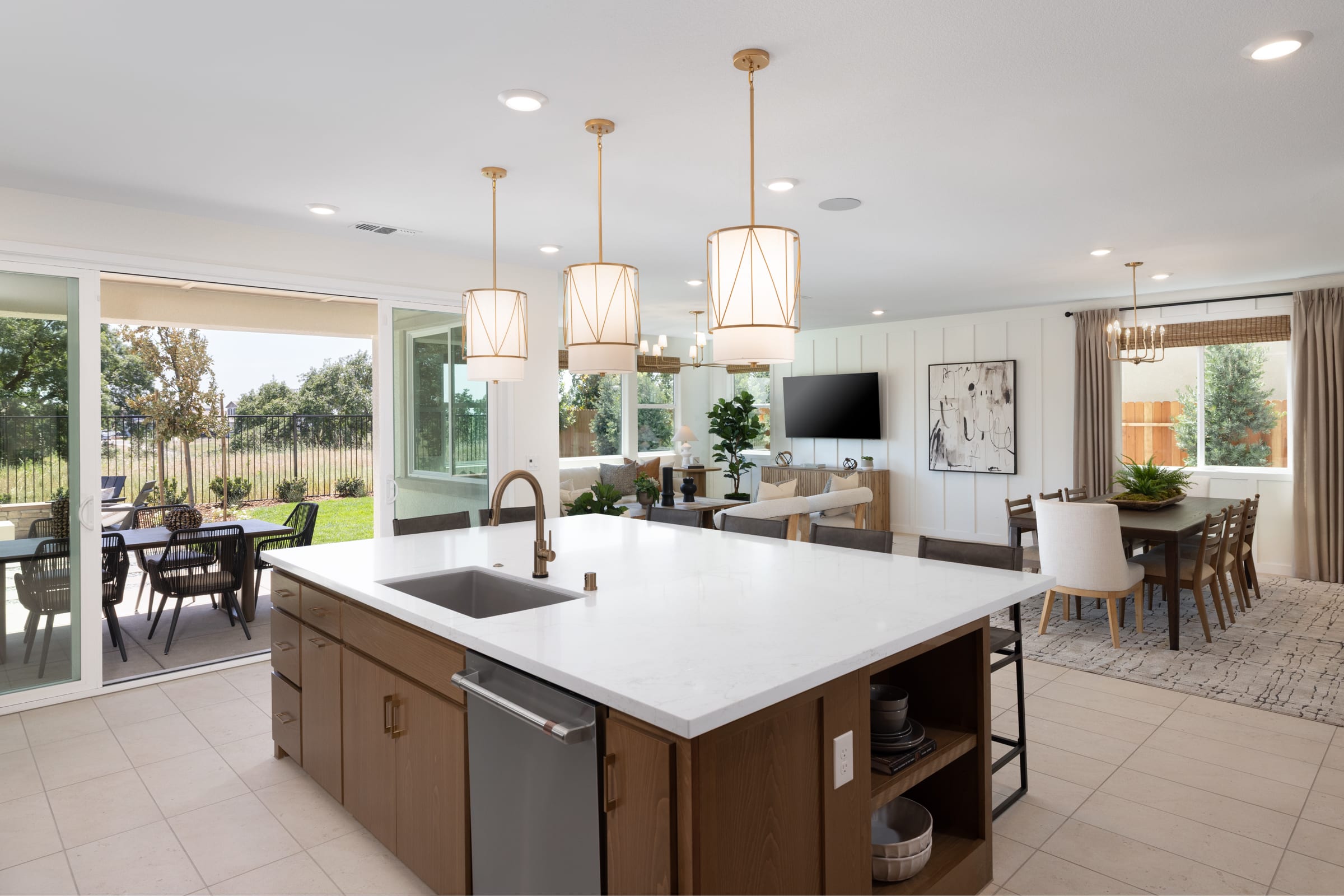 A modern kitchen island with a sink and pendant lights is in the foreground, while a living and dining area with a large television and dining table is visible in the background, opening to a patio with outdoor seating.