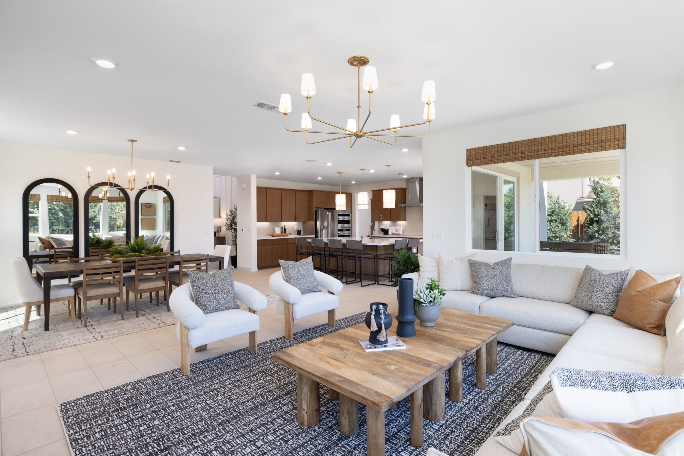 A modern living room with a rustic wooden coffee table and plush white sofas in the foreground, opens to a dining area with arched mirrors and a contemporary kitchen with dark wood cabinets in the background.