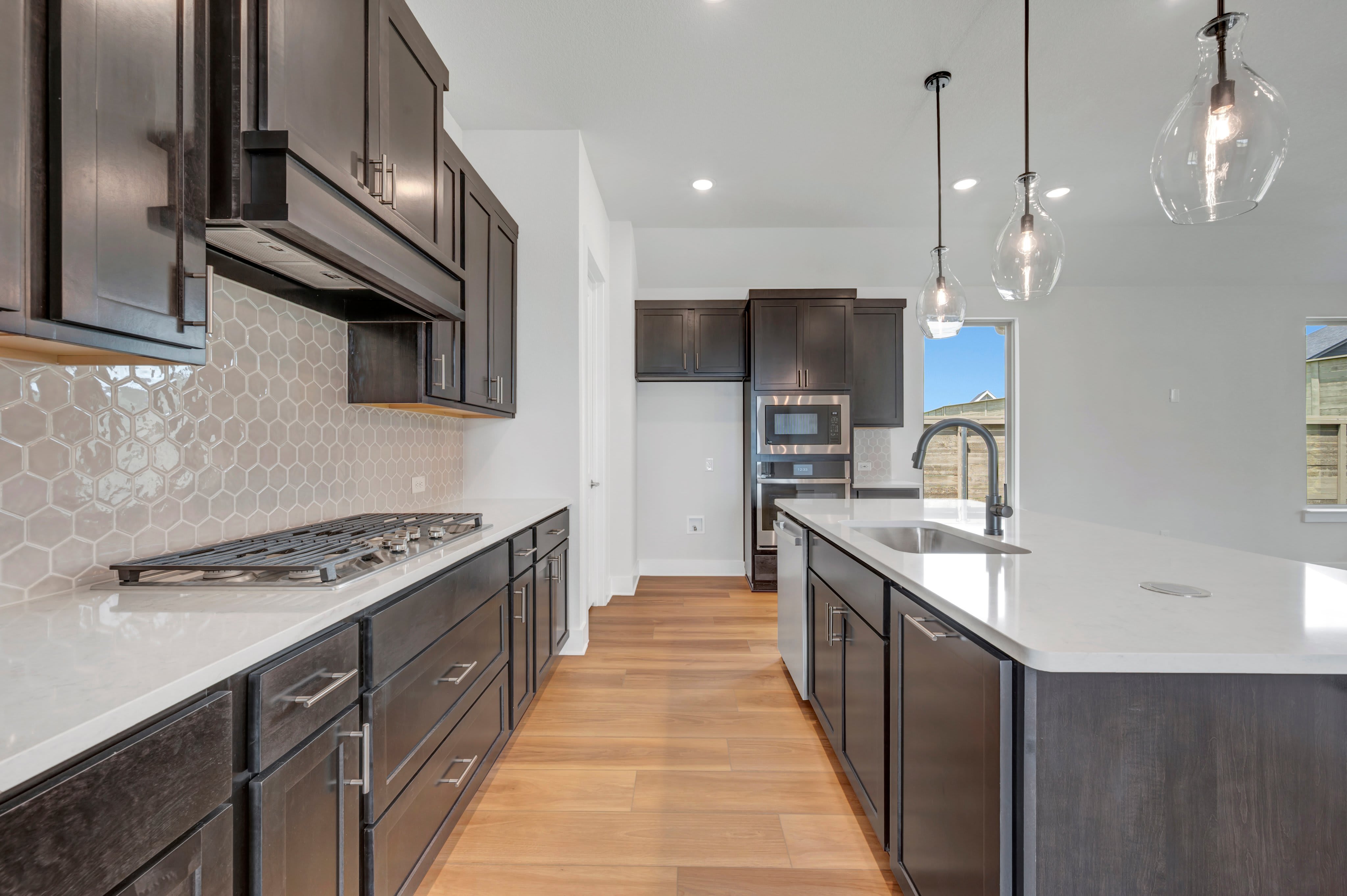 A modern kitchen with dark cabinetry and a white quartz island in the foreground, and stainless steel appliances and a window with a view of a sunny landscape in the background.