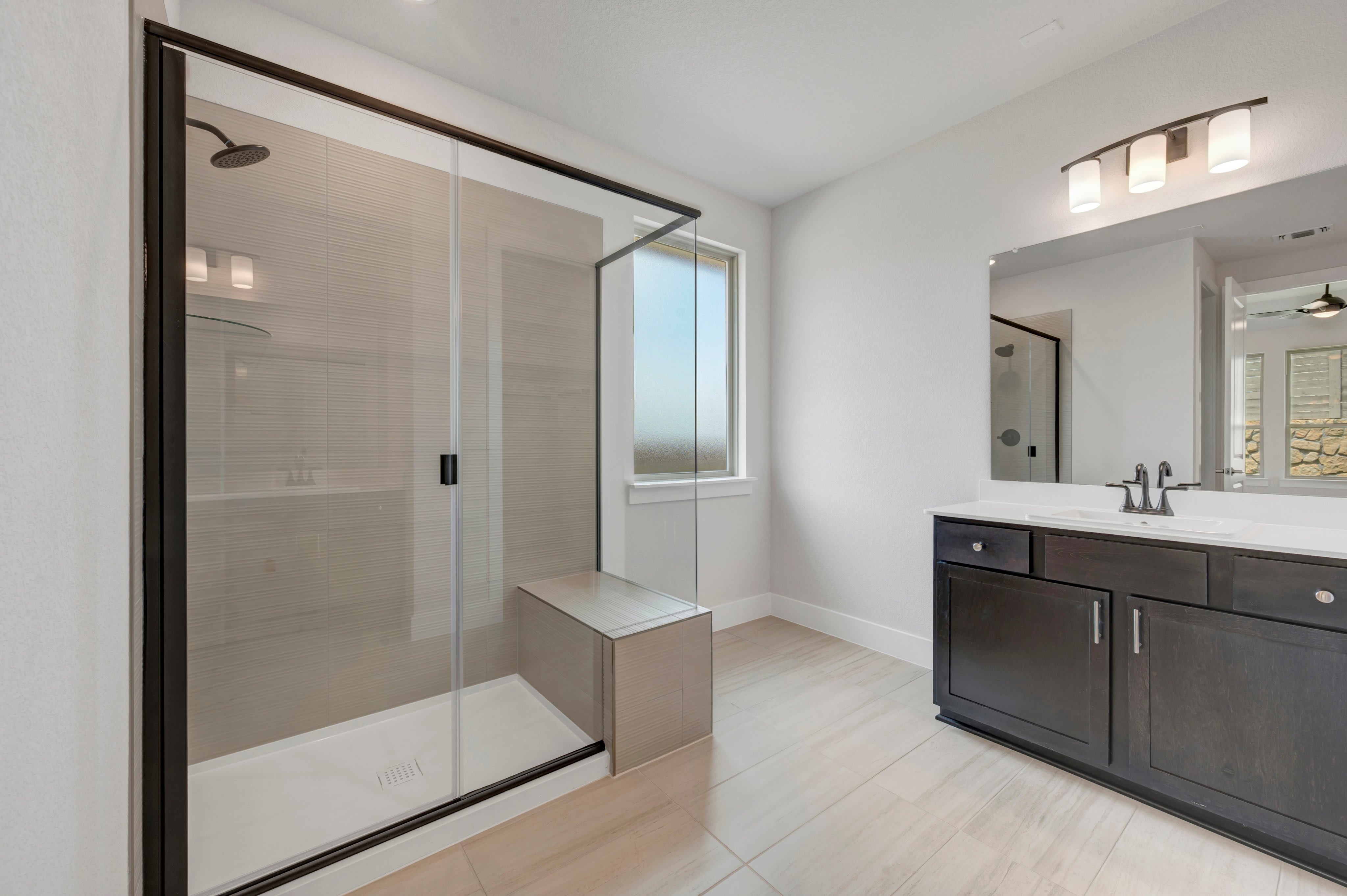 A modern bathroom features a spacious glass-enclosed shower with a built-in bench in the foreground, and a dark wood vanity with a marble countertop and a large mirror in the background.