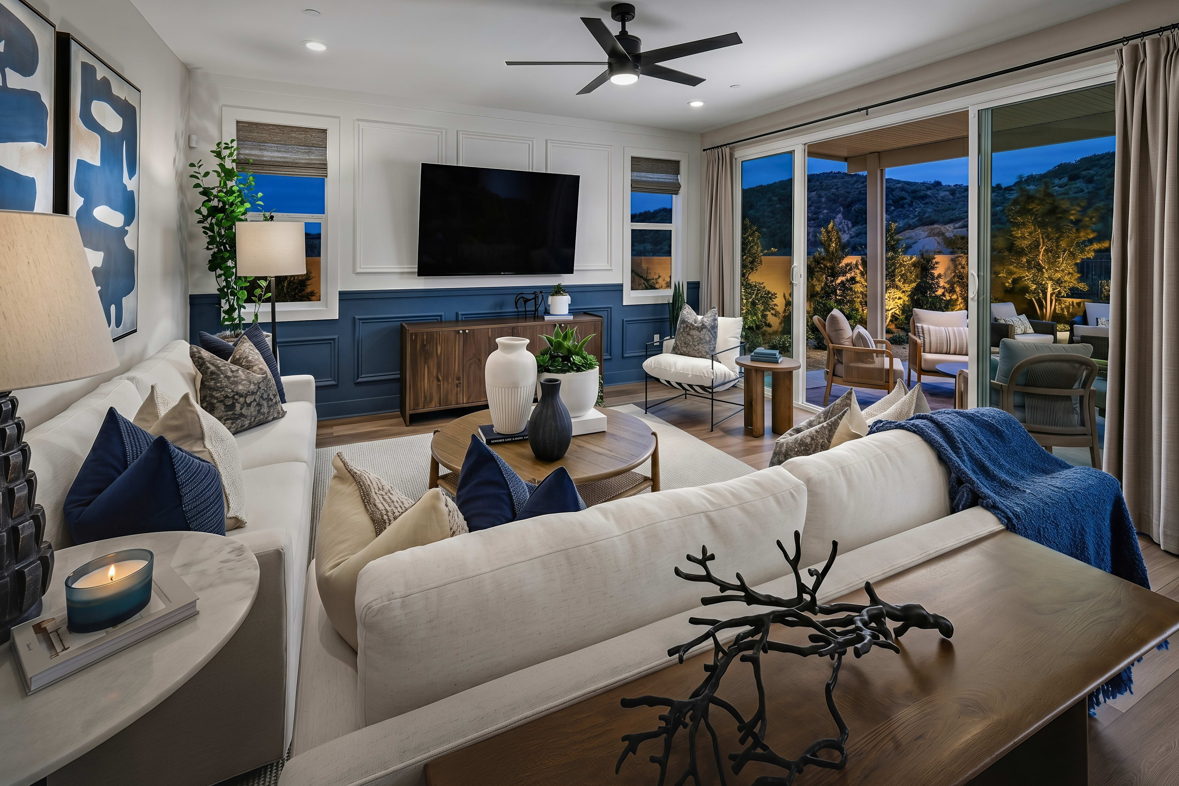 A modern living room with a white sectional sofa and decorative pillows in the foreground, and a patio with seating and a scenic view of hills at dusk in the background.