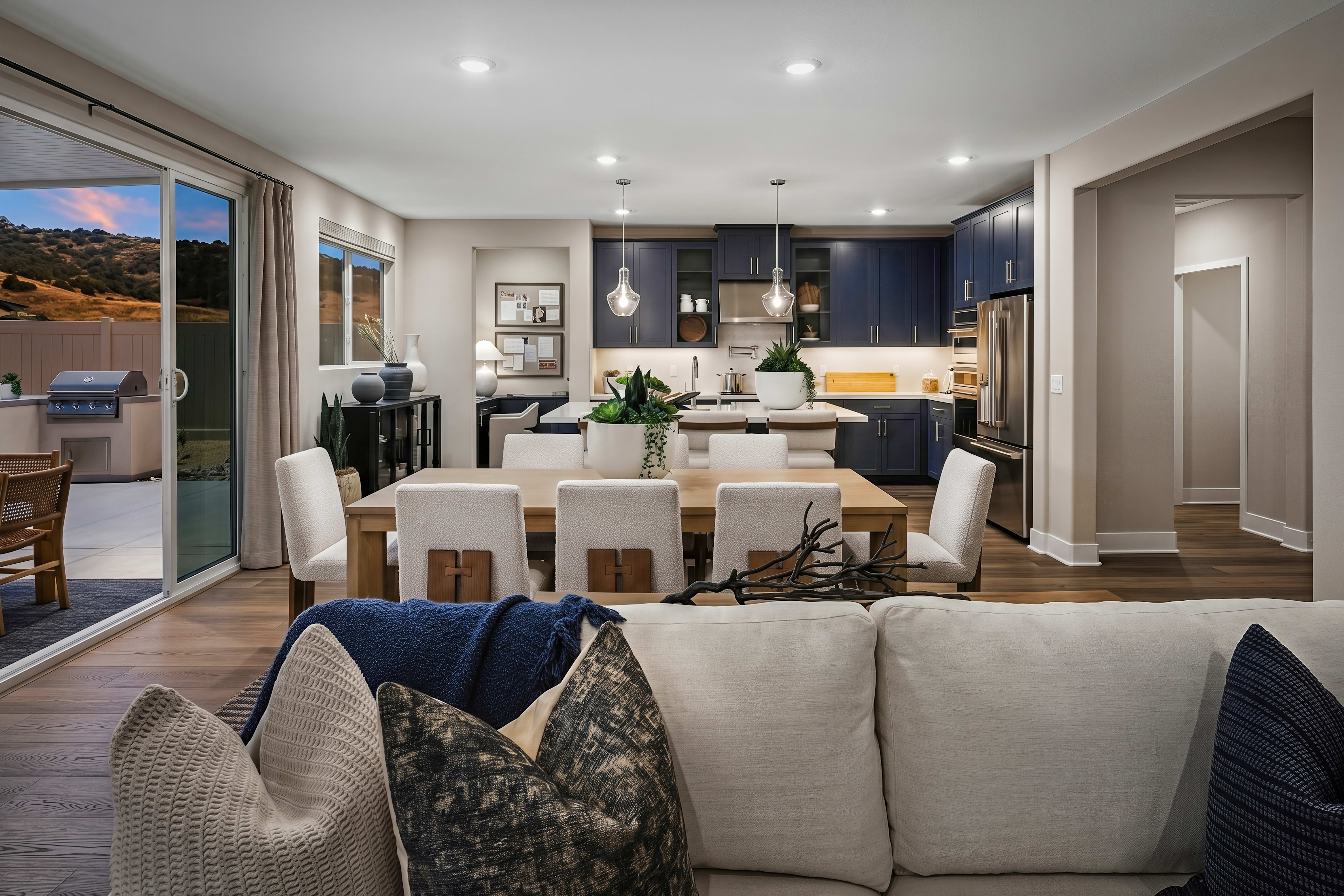 A modern living room with a plush sofa in the foreground opens to a spacious dining area and a navy blue kitchen with stainless steel appliances in the background, beyond which a patio with an outdoor grill is visible through sliding glass doors.
