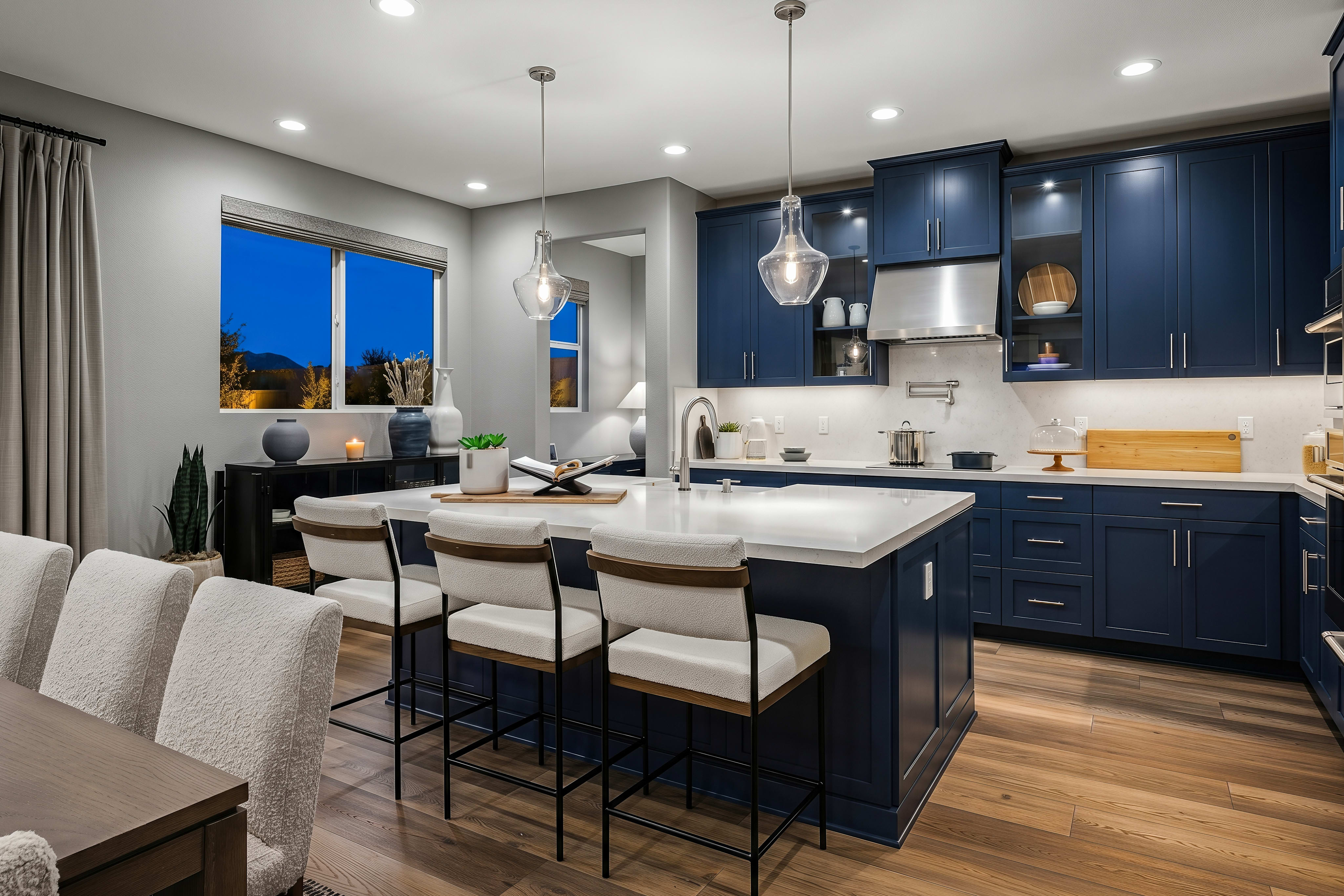 A modern kitchen with a blue island and white stools in the foreground is set against a backdrop of dark blue cabinets and a window showing a twilight sky.
