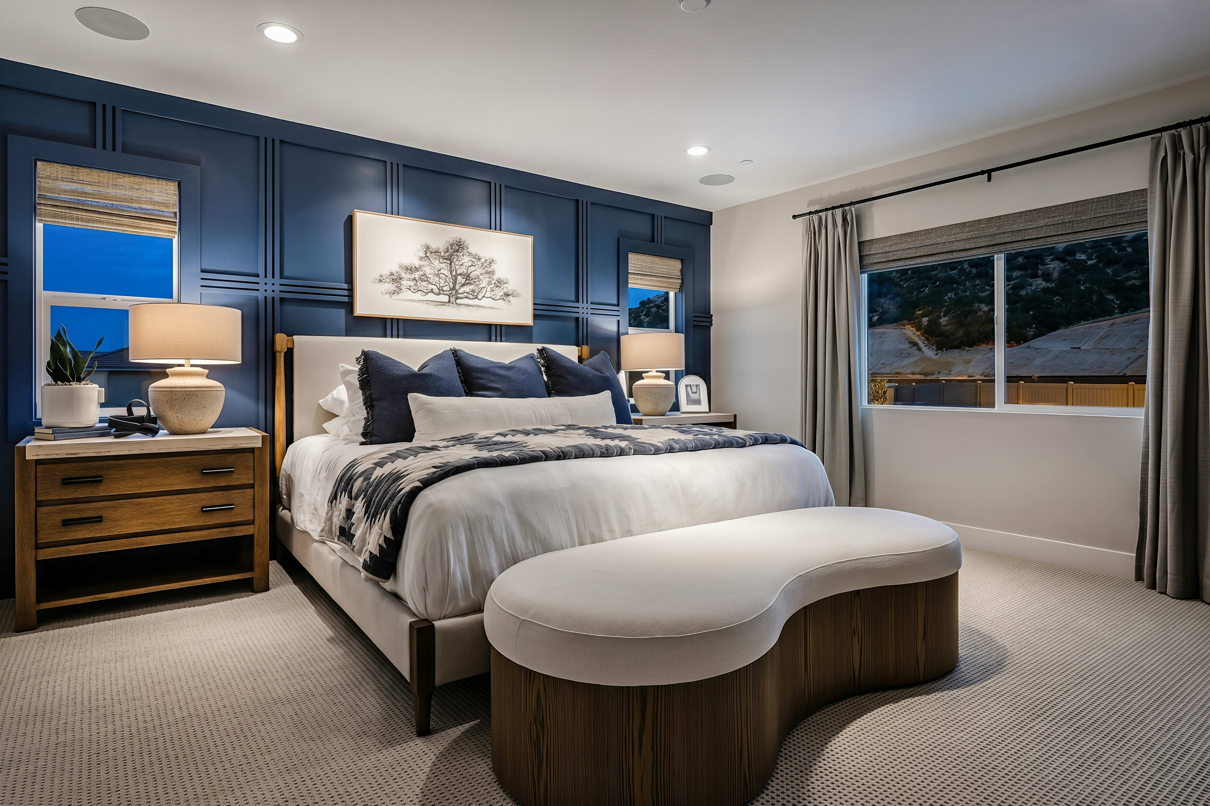 A luxurious bedroom with a plush bed and a curved bench in the foreground is set against a dark blue paneled wall with windows and a framed artwork in the background.