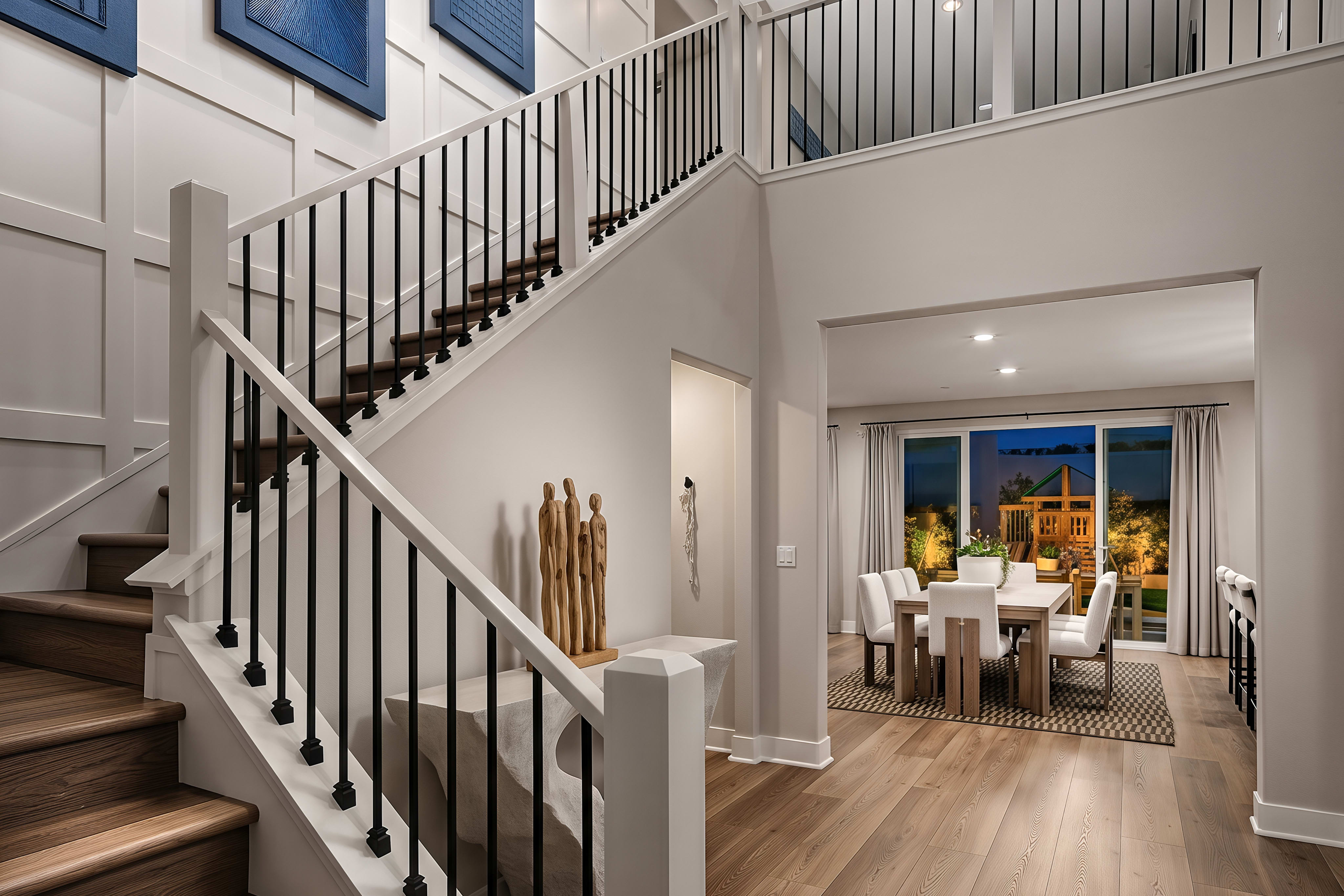 A modern staircase with wooden steps and black railings leads up to a loft overlooking a dining room with a large table and chairs, all set against a backdrop of contemporary wall art.