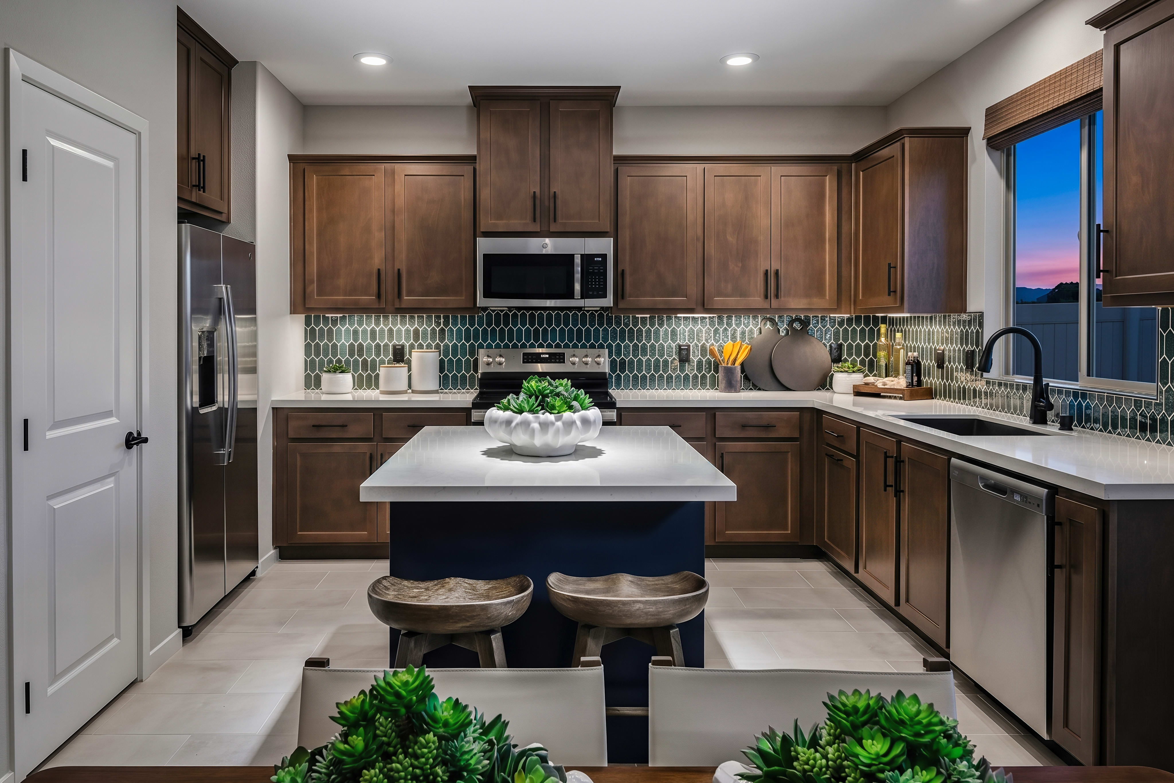 A modern kitchen with a dark blue island and stools in the foreground, and dark wood cabinets and a tiled backsplash in the background.