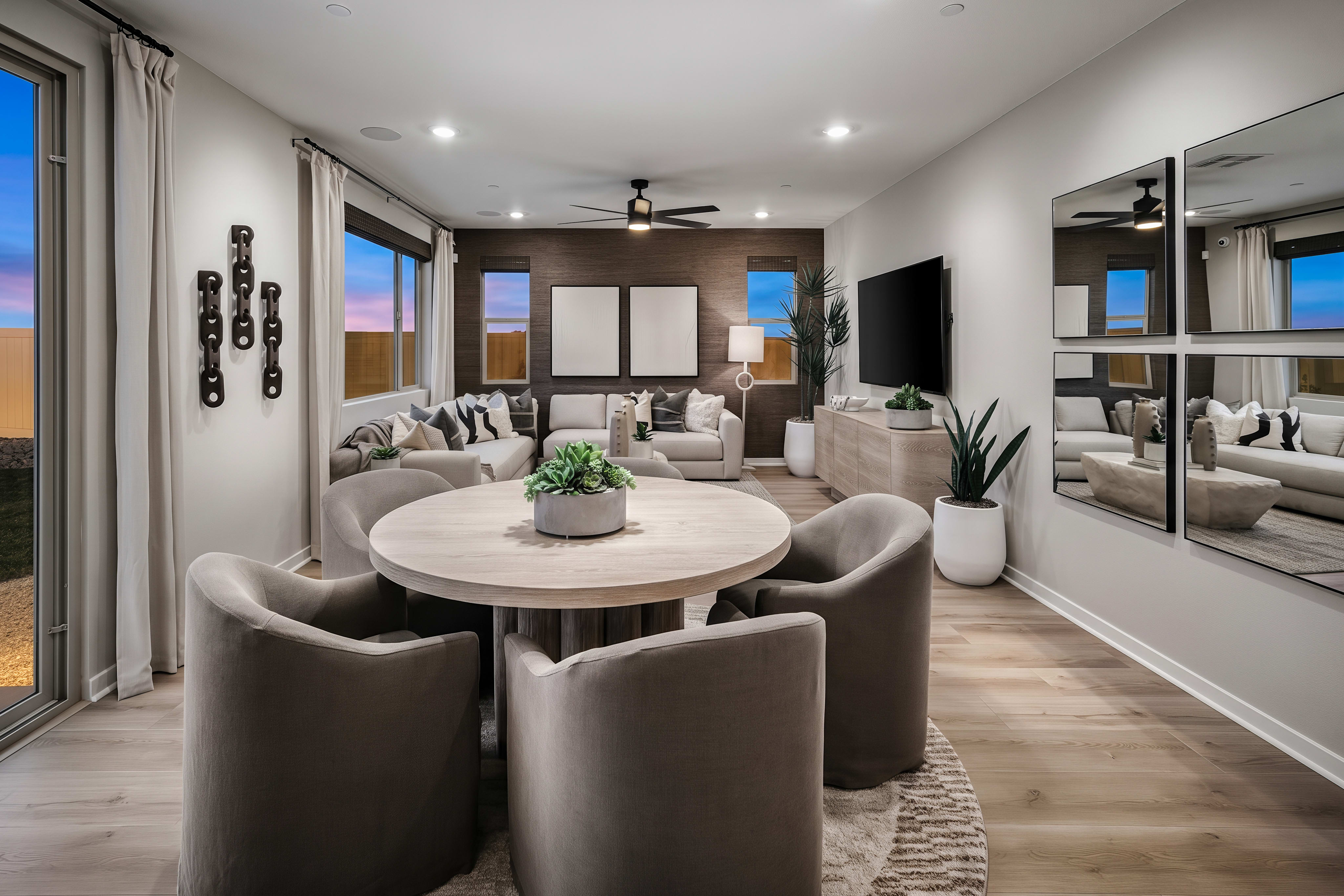 A round wooden dining table with four plush chairs sits in the foreground, while a spacious living room with a television and seating area is visible in the background.