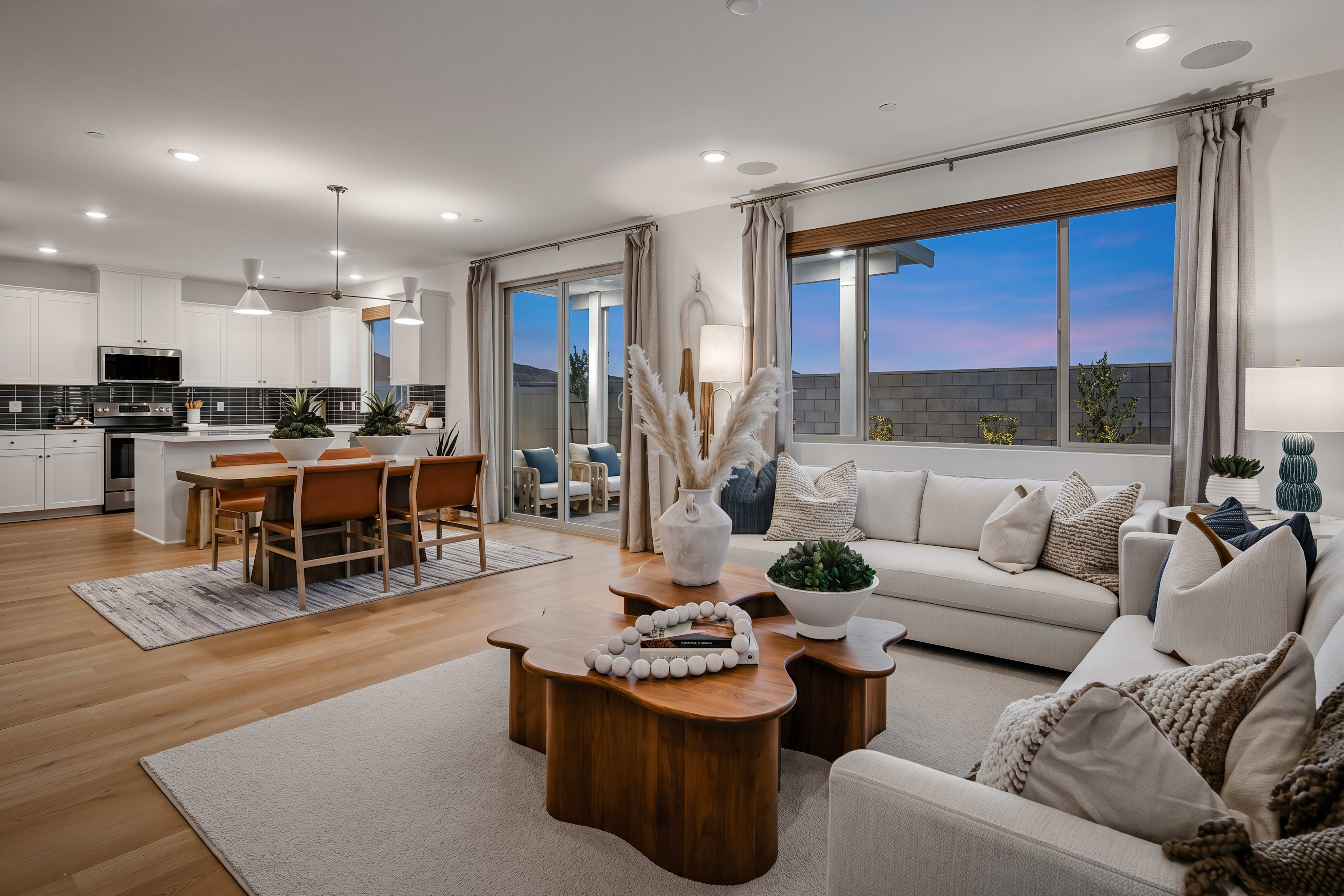 A modern living room with a large sectional sofa and a unique wooden coffee table in the foreground, opens to a dining area and kitchen, with large windows revealing a twilight sky in the background.