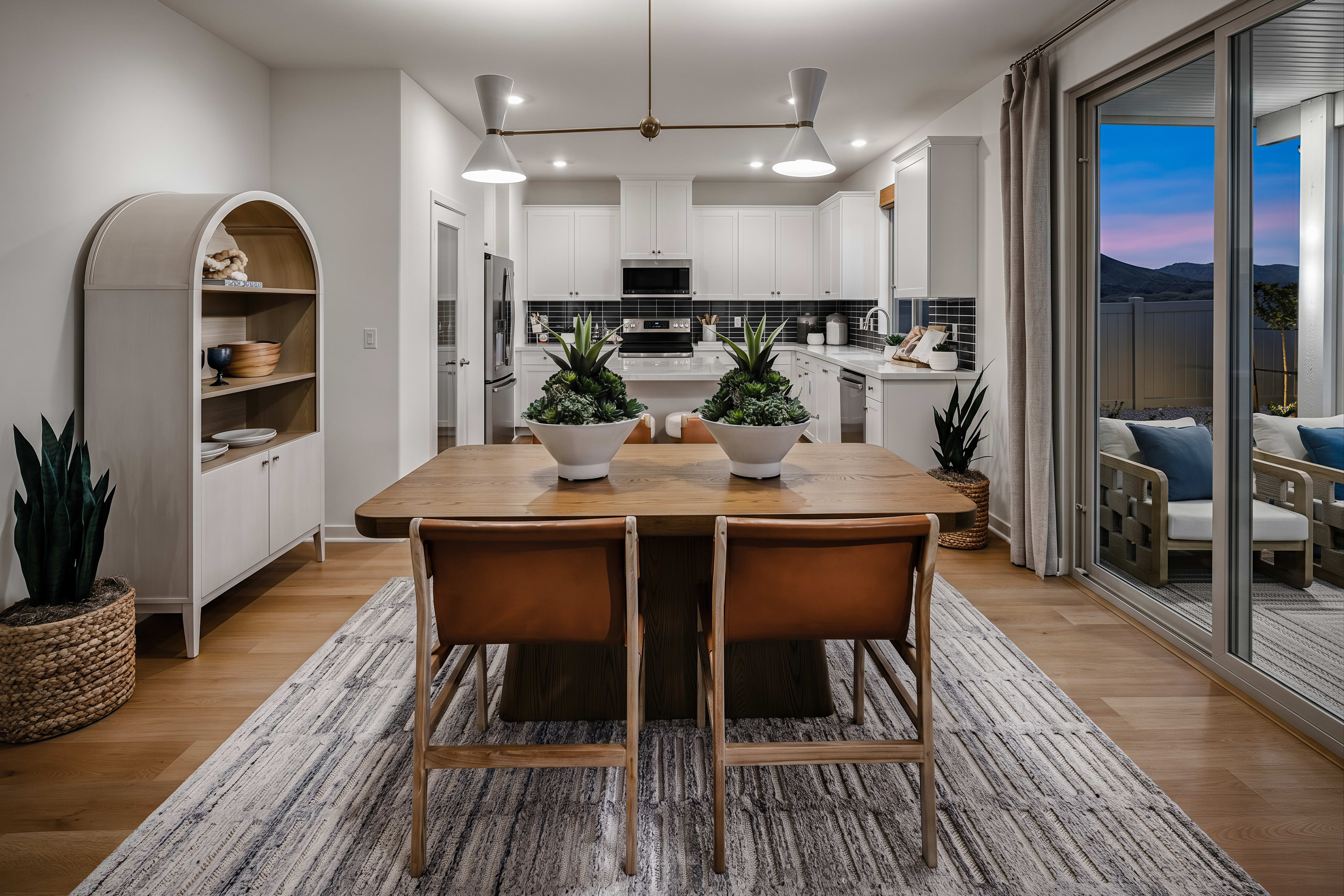 A wooden dining table with two chairs sits on a patterned rug in the foreground, while a modern kitchen with white cabinets and a tiled backsplash is visible in the background.