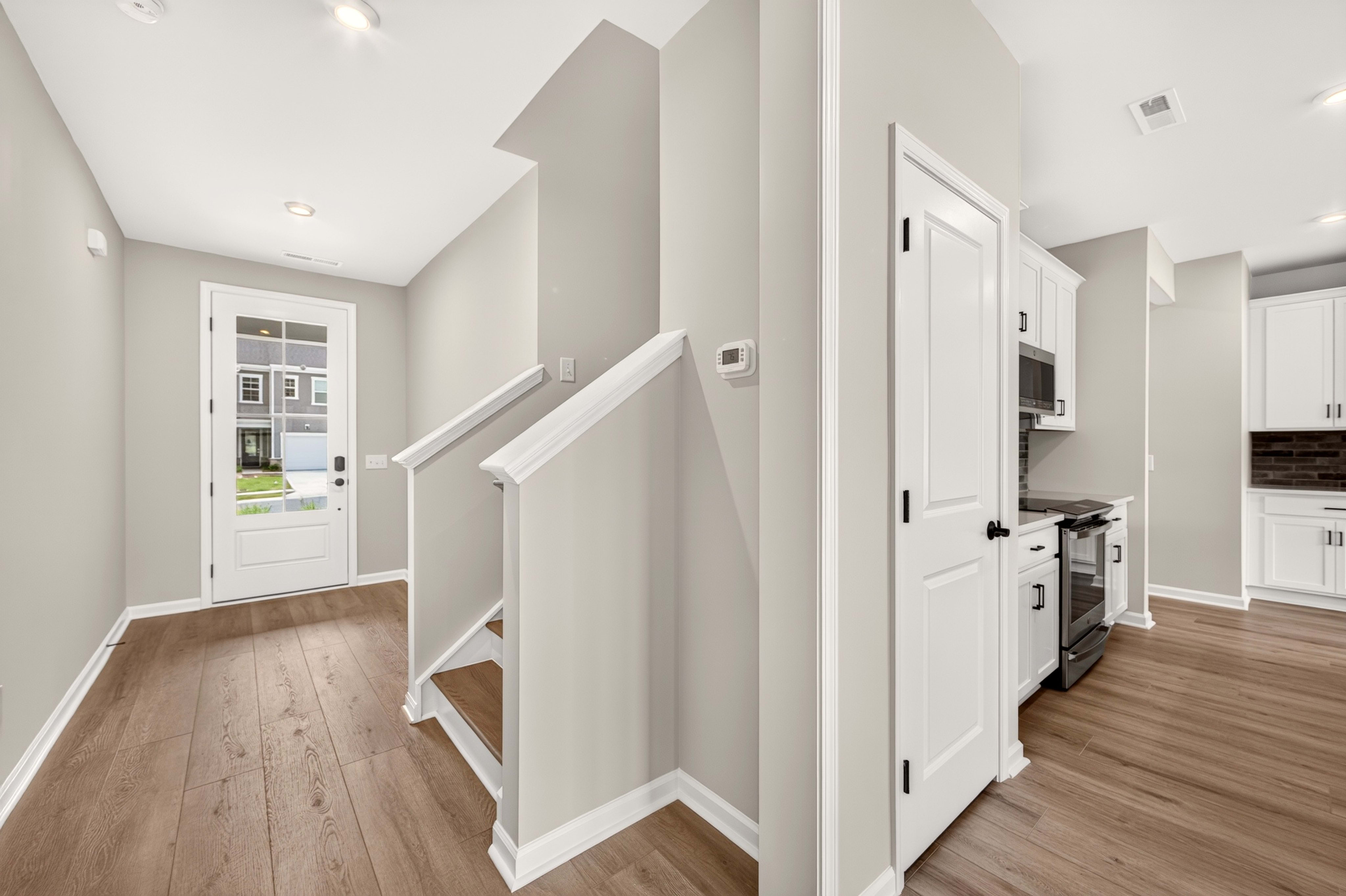 A modern interior with a wooden floor in the foreground leads to a white staircase and a doorway, with a kitchen visible in the background.