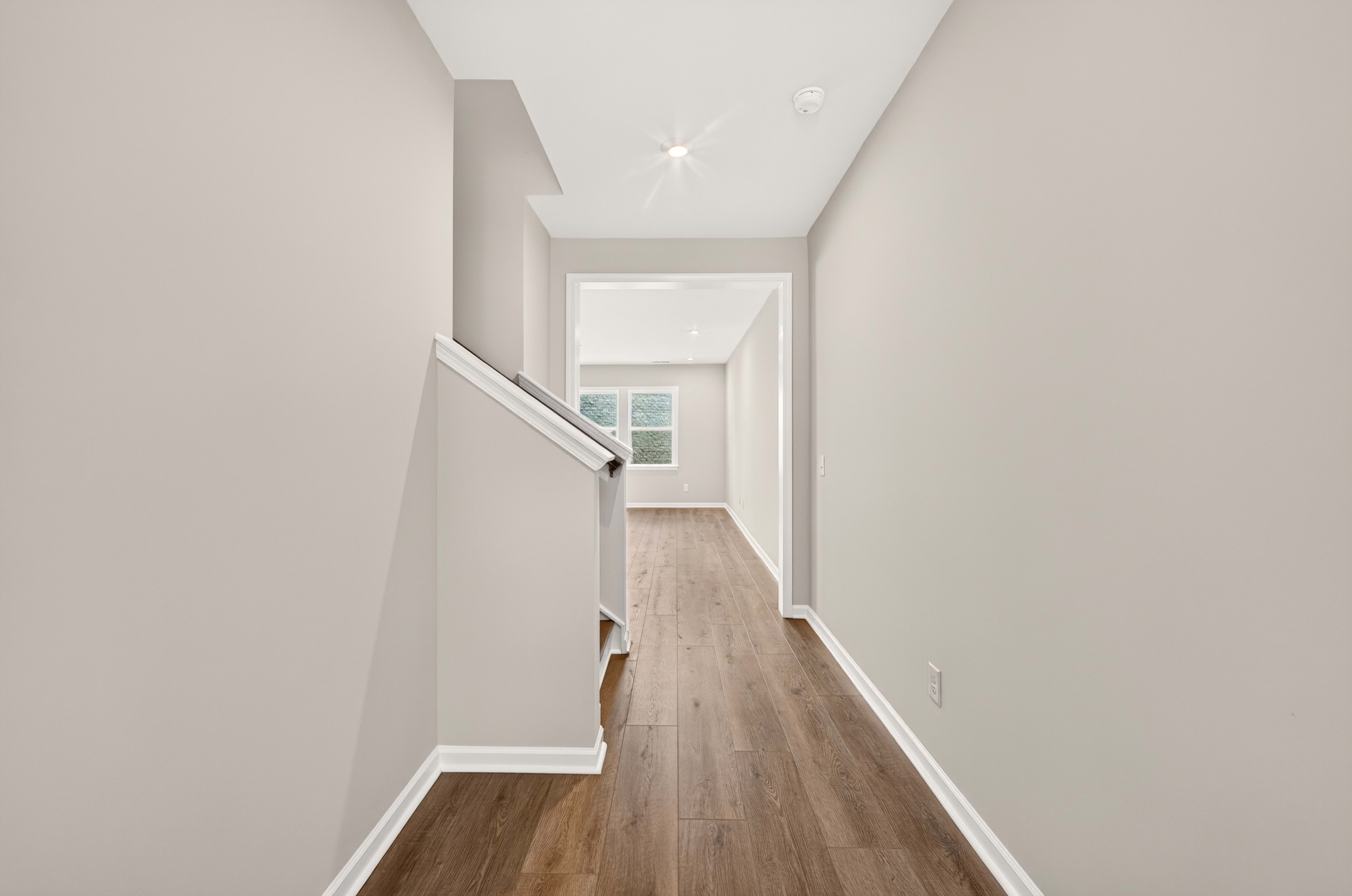 A light-colored hallway with wood-look flooring leads to a bright, open room with large windows.