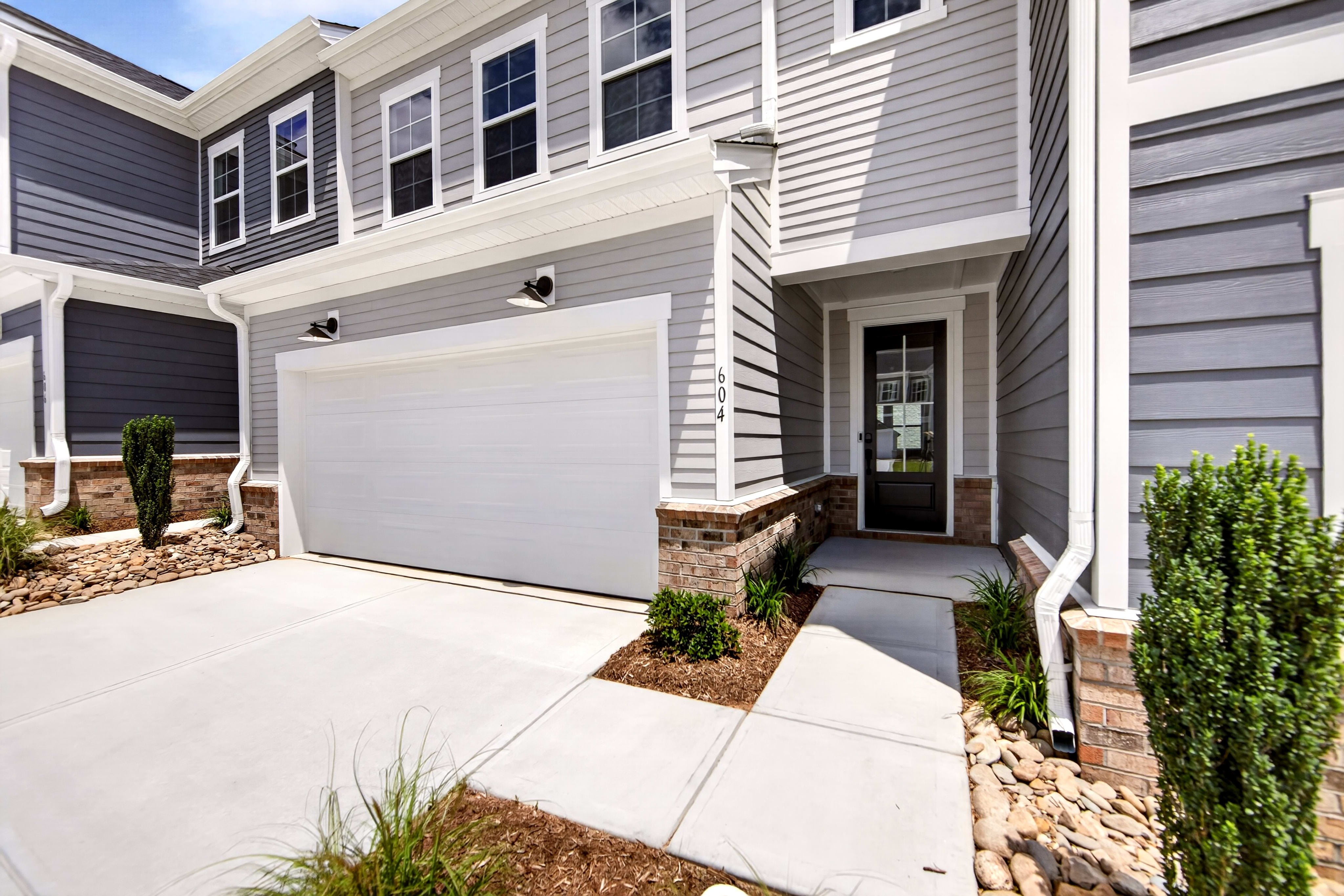 A modern townhouse with a white garage door and a glass-paneled front door is set against a backdrop of gray siding and a bright blue sky.
