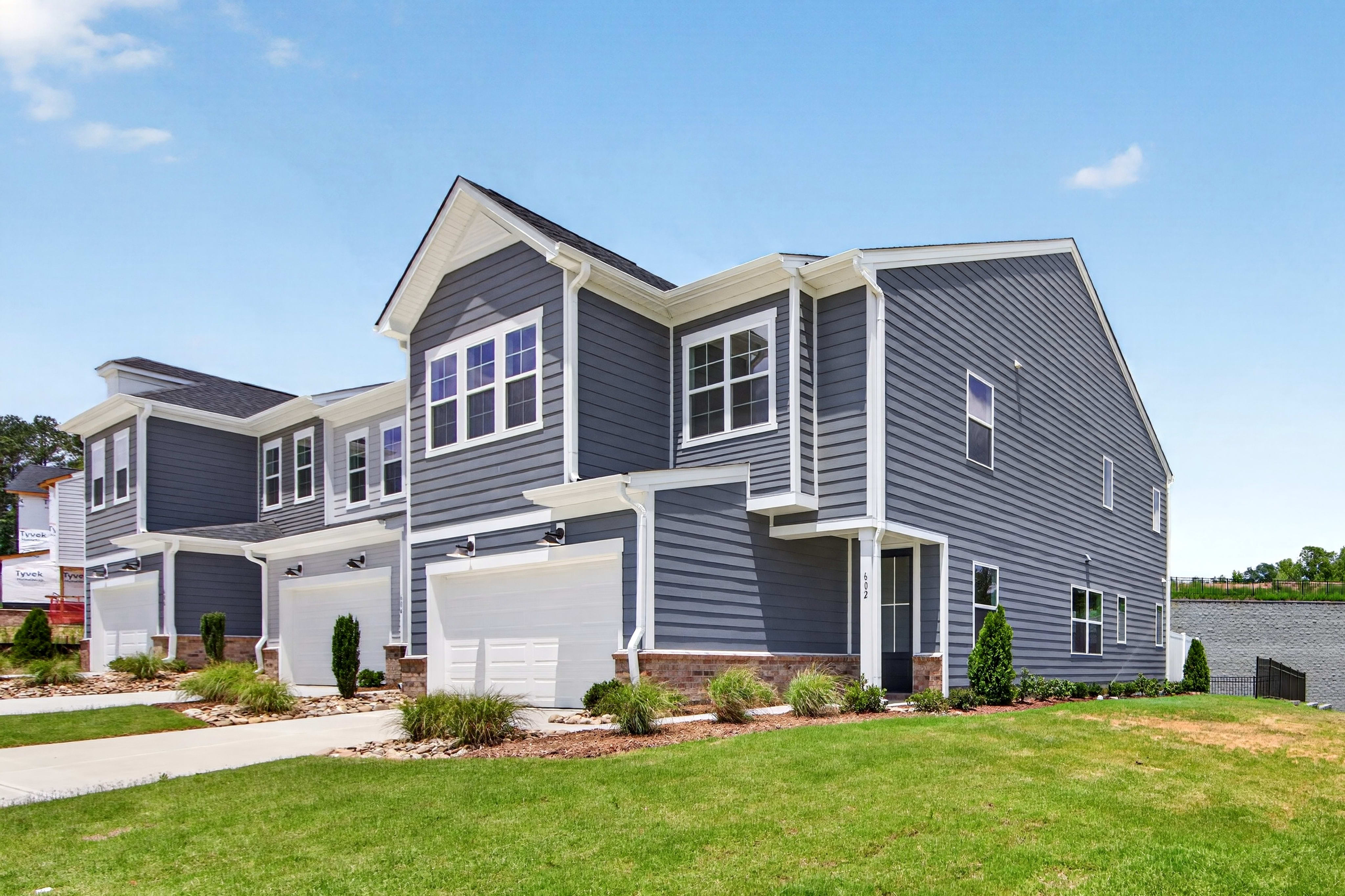 A row of modern townhouses with gray siding and white trim stands on a manicured lawn under a clear blue sky.