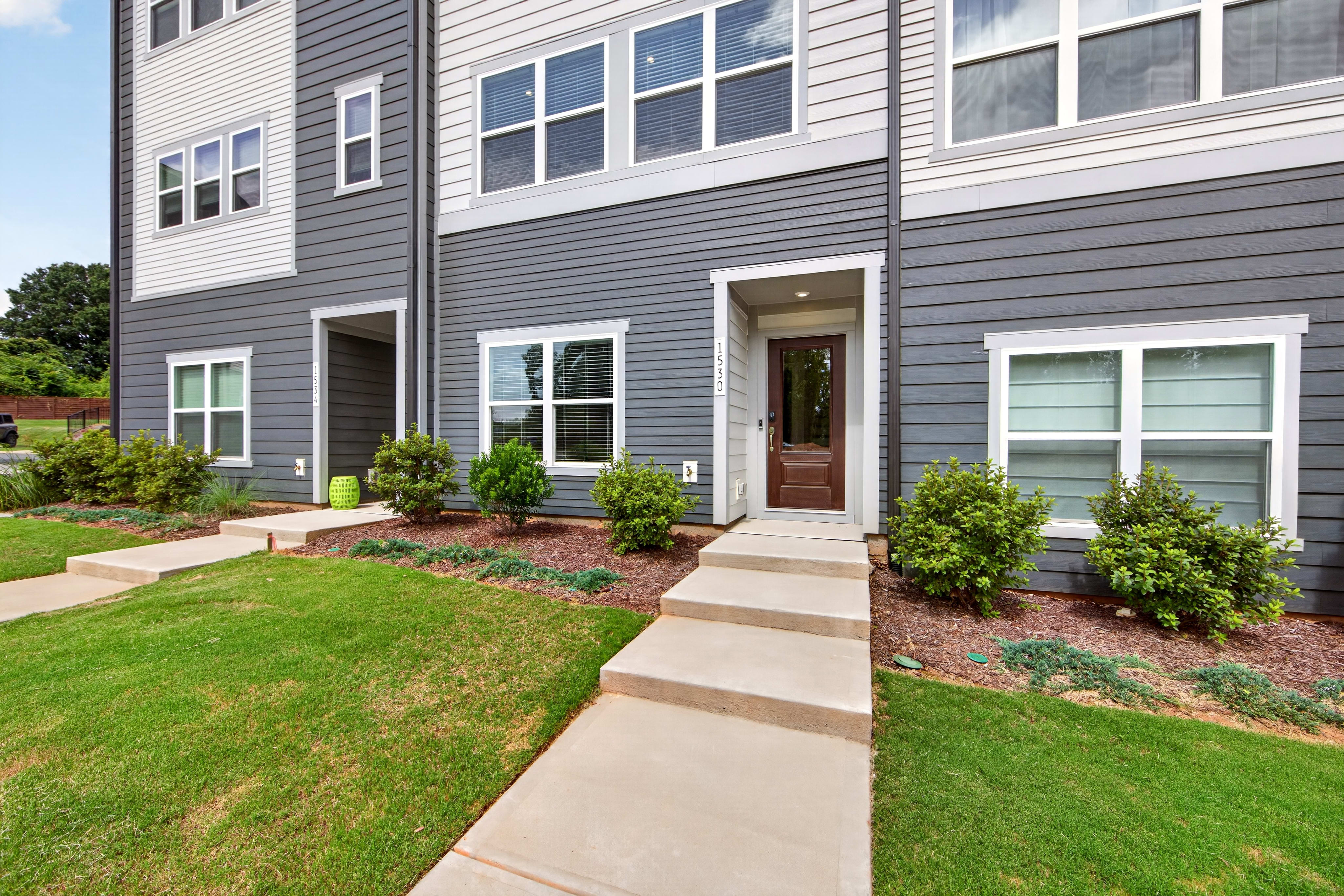 A concrete walkway leads through a green lawn and manicured bushes to the entrance of a modern gray and white townhouse with large windows.