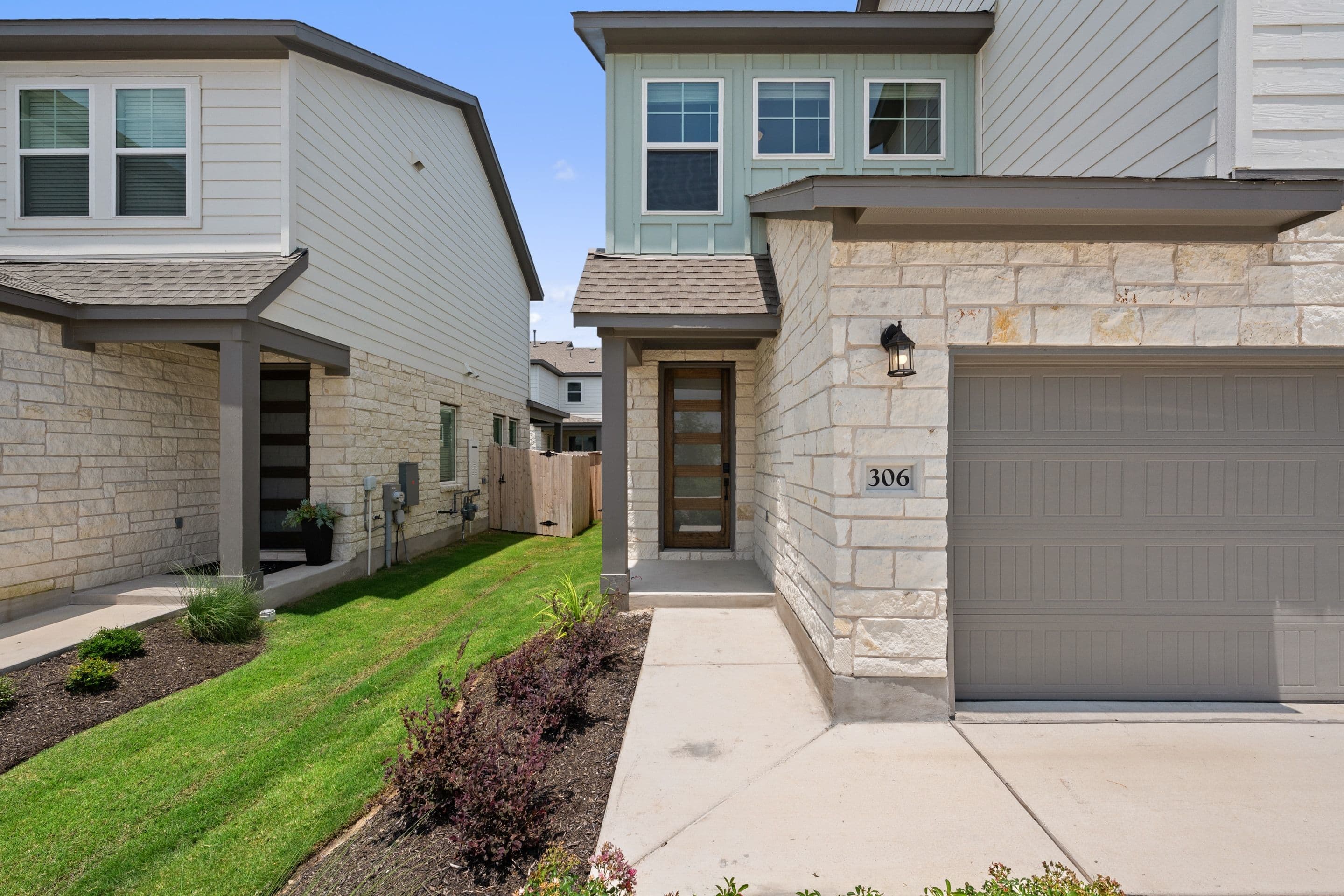 A modern townhouse with a stone facade and a gray garage door stands on a concrete walkway, with a lush green lawn and landscaping in the foreground, and other similar townhouses in the background.