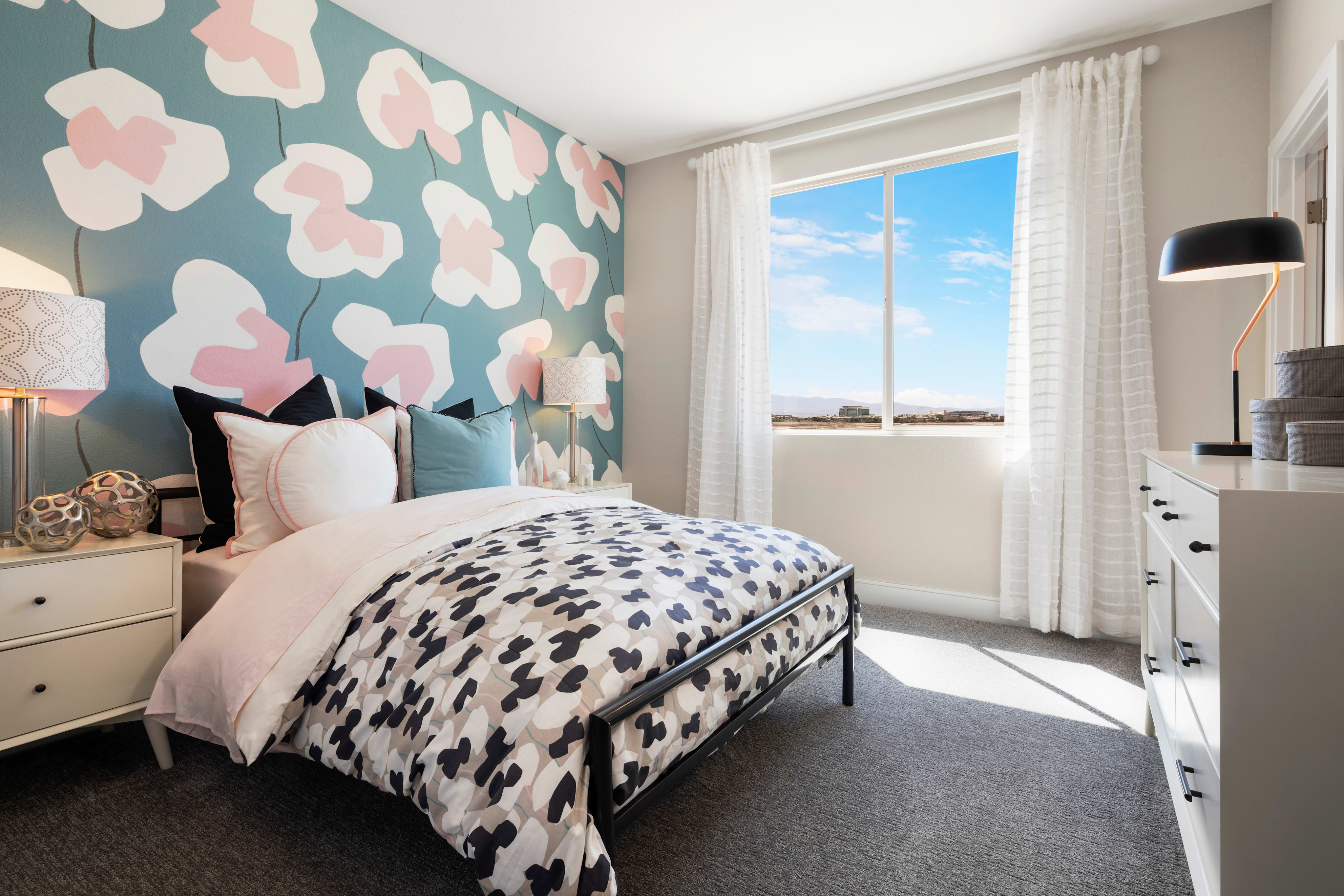 A bedroom with a patterned duvet and floral wallpaper in the foreground is bathed in sunlight from a large window revealing a bright, cloudy sky and distant landscape in the background.