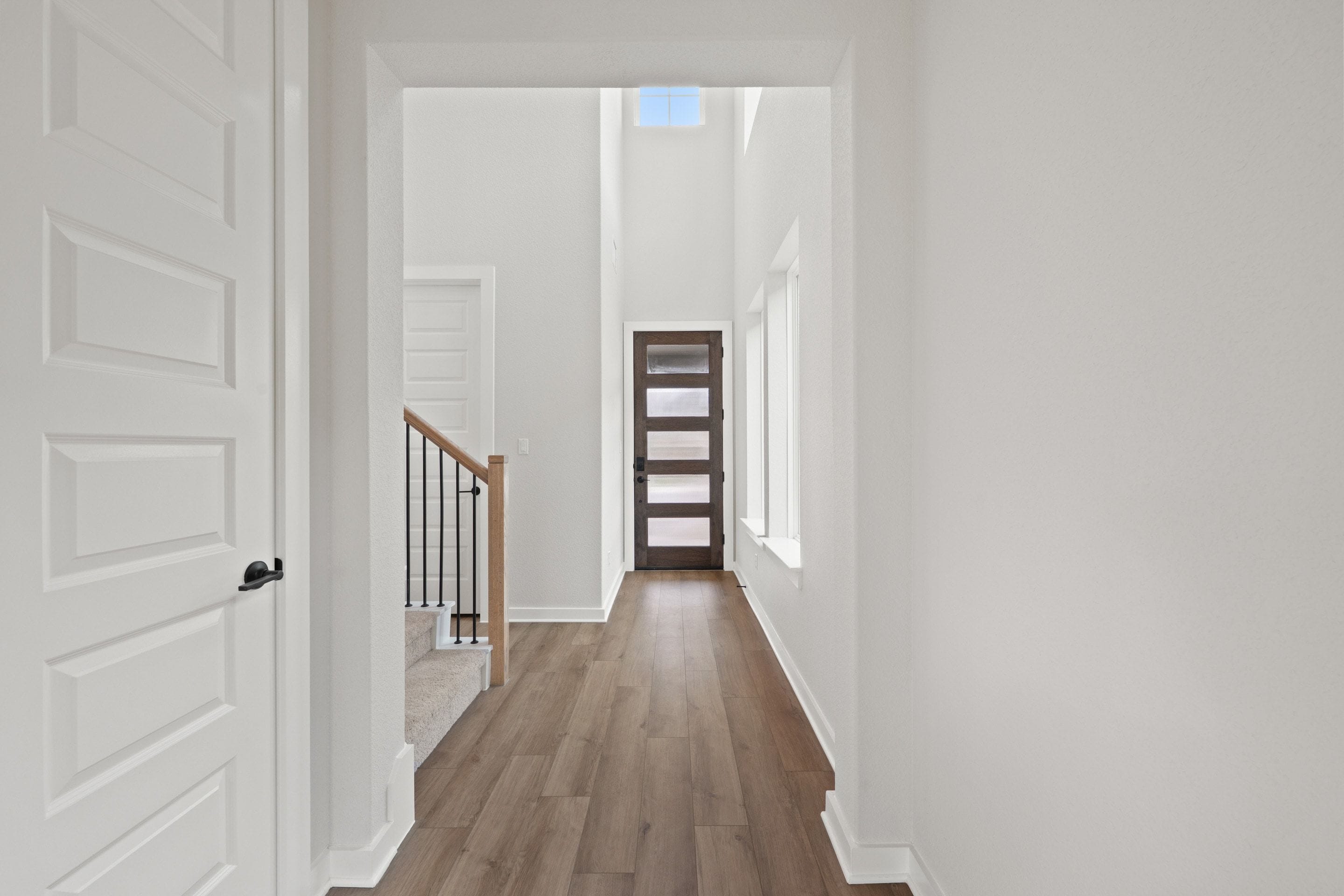 A modern entryway with a dark wood door and frosted glass panels is visible at the end of a hallway with wood-look flooring, while a white door and staircase with a metal railing are in the foreground.