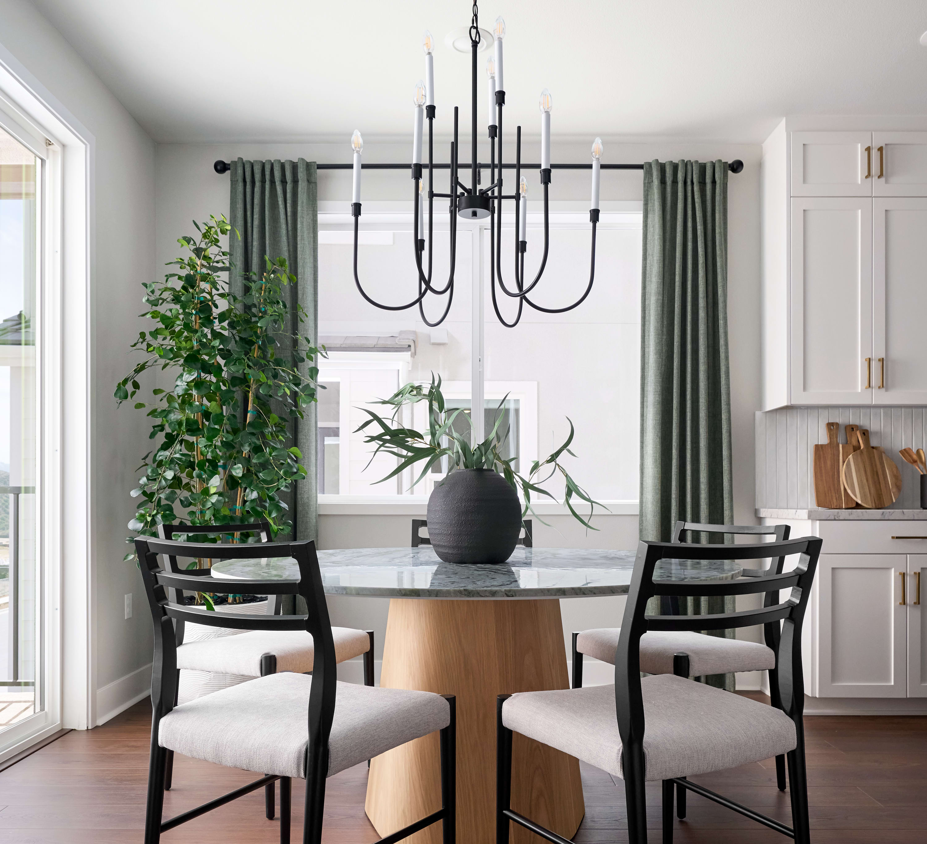 A modern dining set with upholstered chairs surrounds a round table with a vase of greenery in the foreground, while a large window with green curtains and kitchen cabinets form the background.