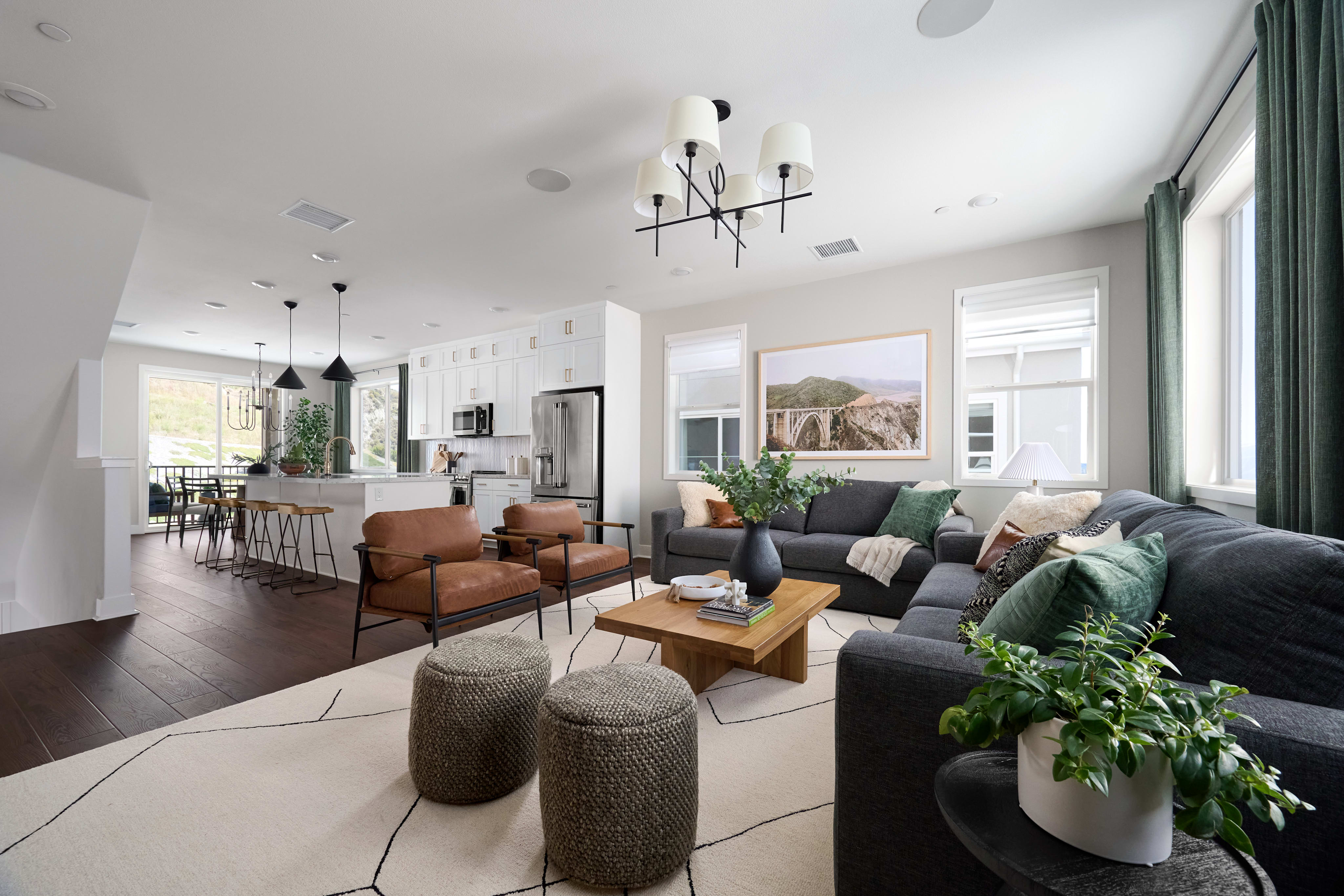 A modern living room with a dark sectional sofa and two leather armchairs in the foreground is adjacent to a bright, open-plan kitchen with white cabinets and stainless steel appliances in the background.