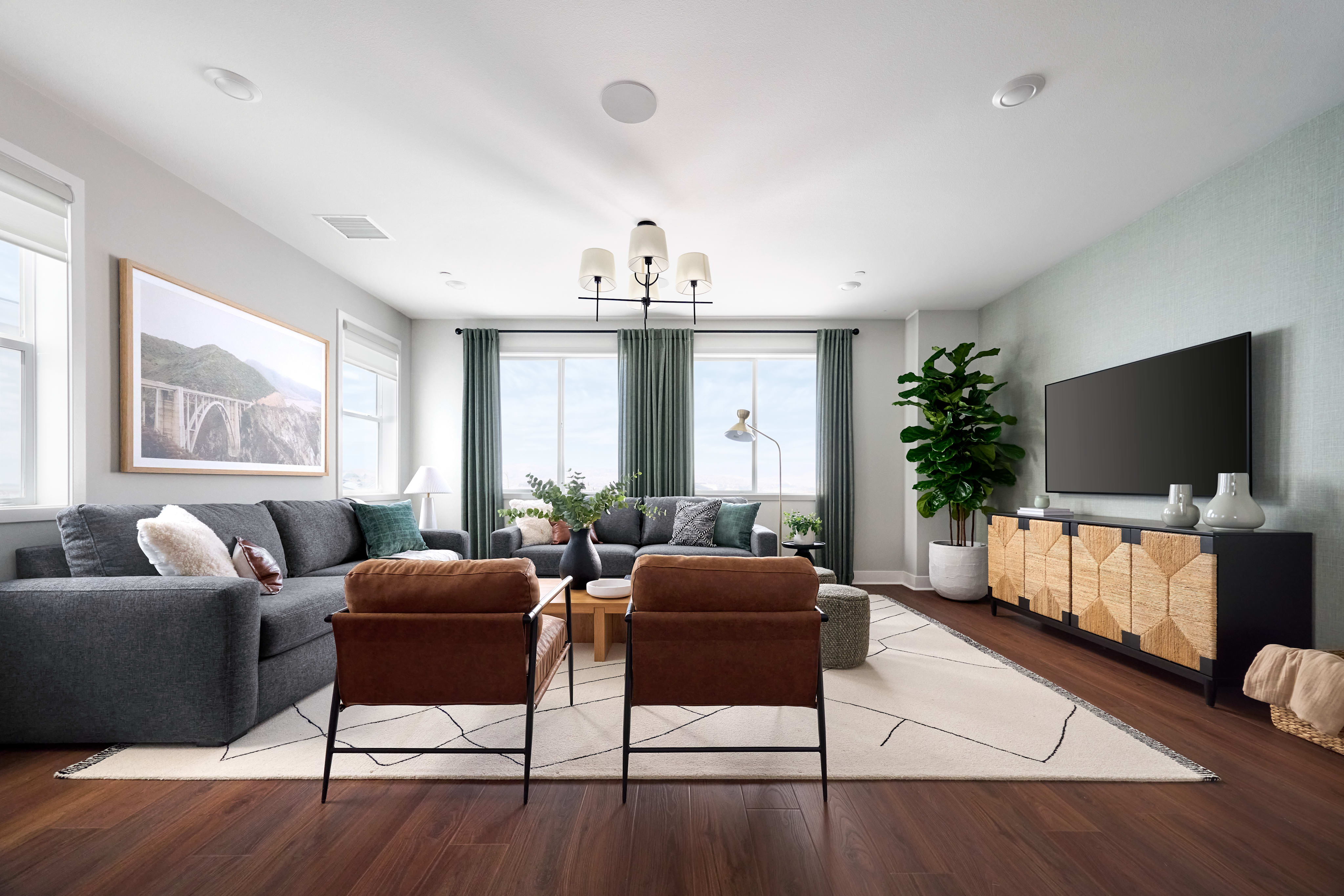 Two brown leather armchairs face a large sectional sofa in the foreground, while a television and a large plant are visible in the background.