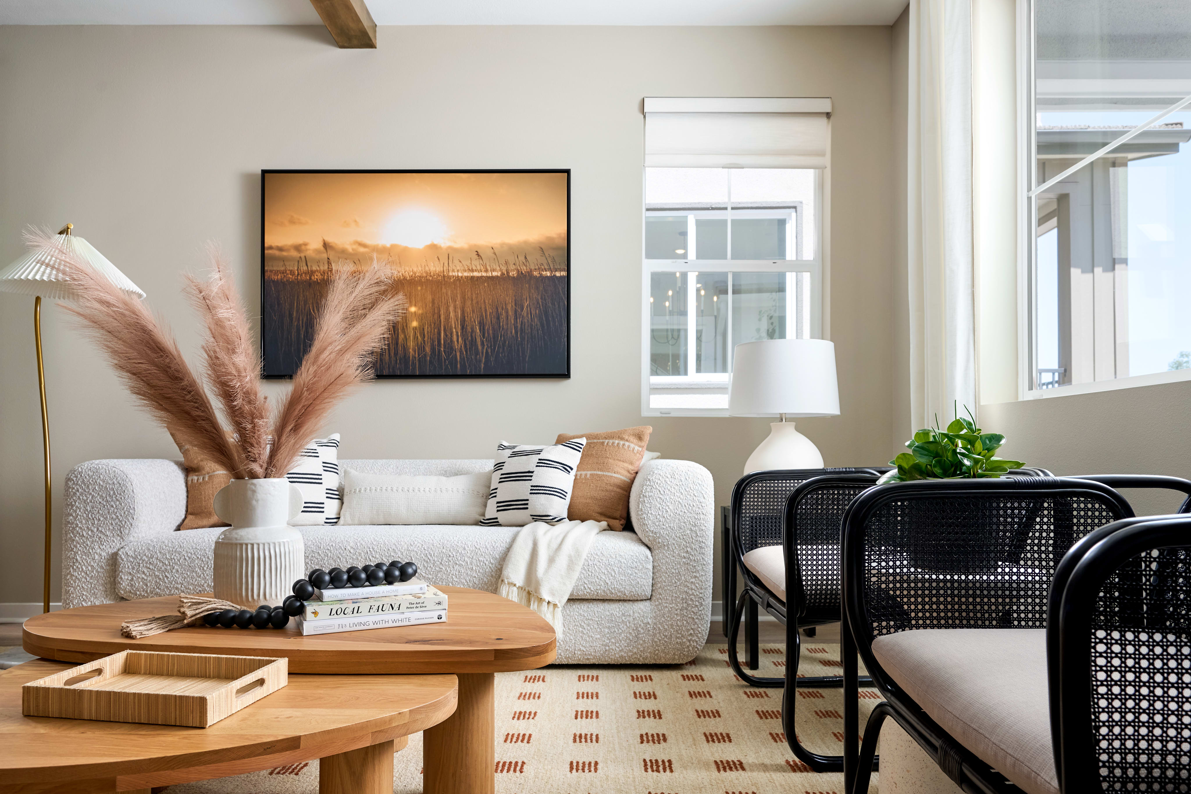 A cozy living room features a white boucle sofa with decorative pillows and a large landscape painting above it, while two black rattan chairs and a wooden coffee table with a tray of decorative items fill the foreground.