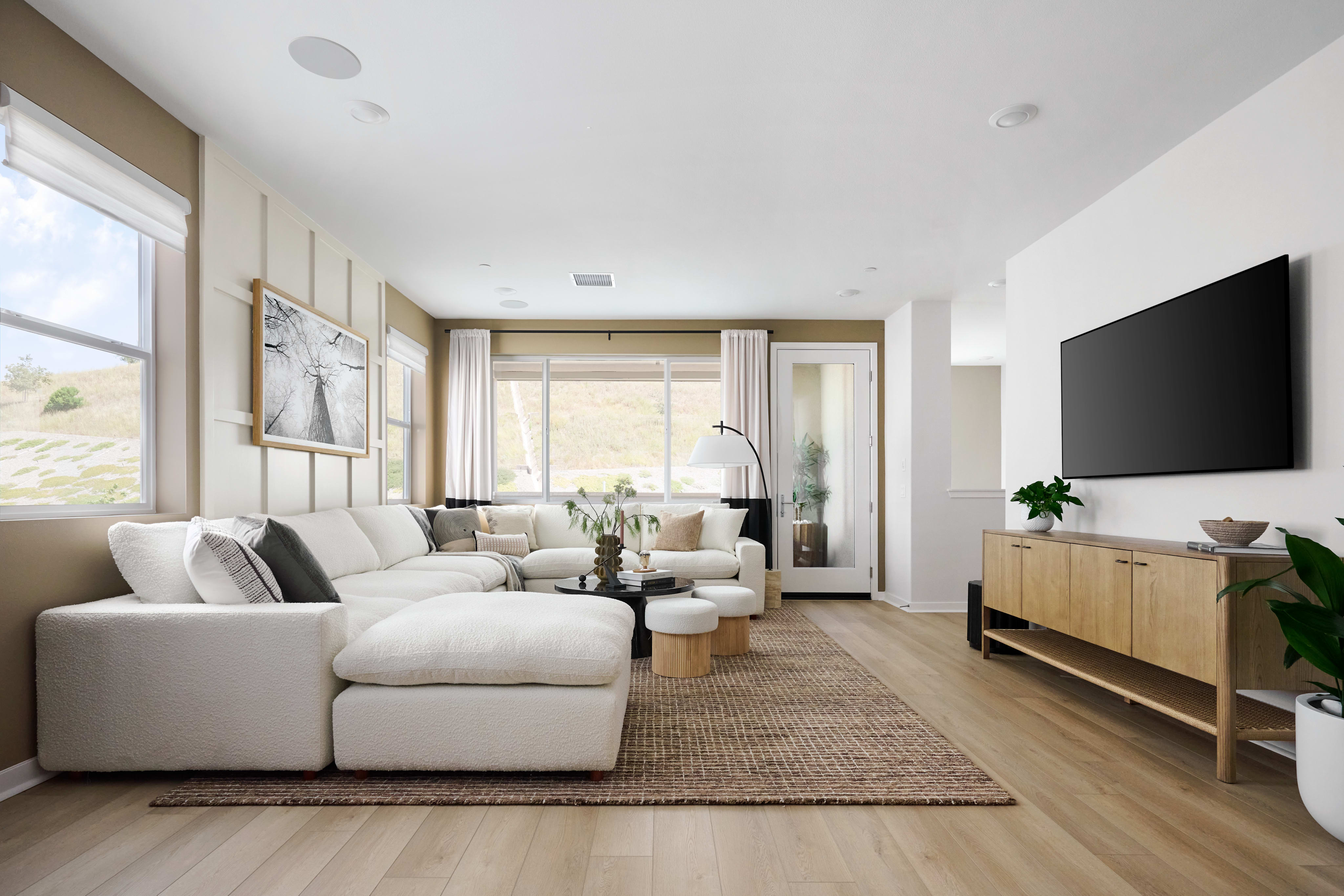 A plush white sectional sofa with striped pillows sits on a woven rug in the foreground, while a large television mounted on a light wood cabinet is visible in the background.