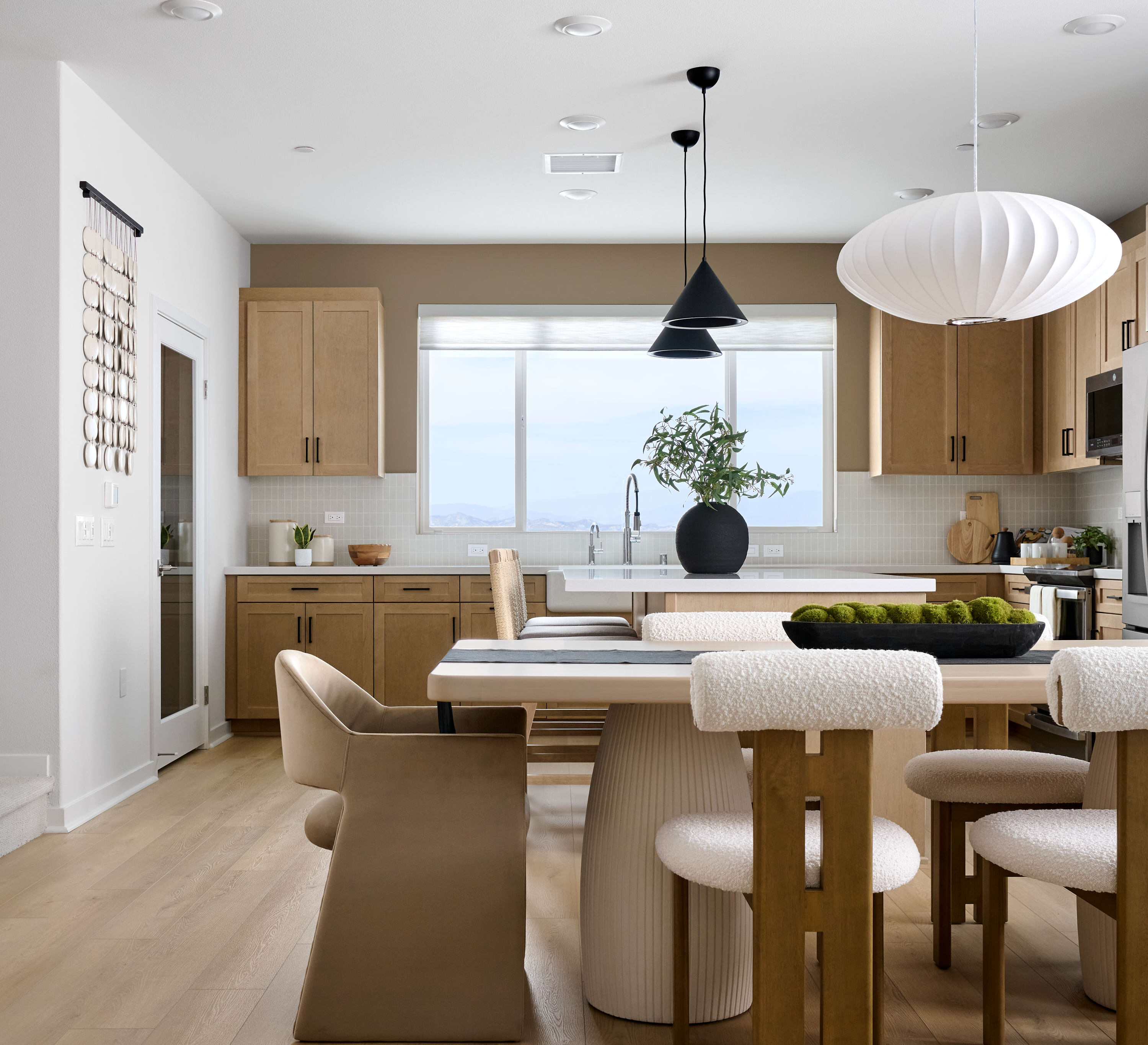A modern dining area with a light wood floor and a large white table with textured chairs in the foreground is set against a bright kitchen with light wood cabinets and a large window in the background.