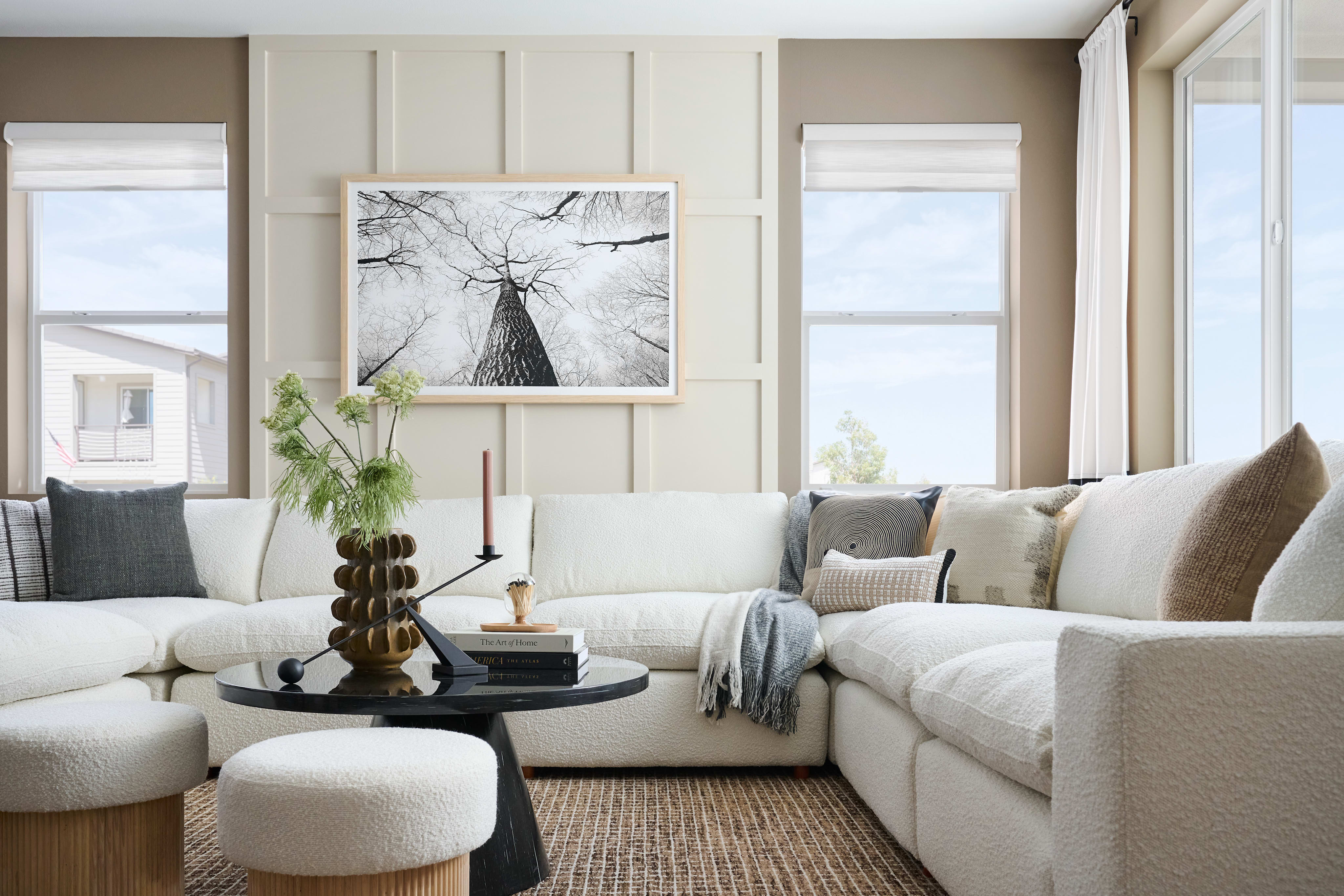 A plush white sectional sofa with decorative pillows sits around a black coffee table in the foreground, while a framed black and white tree canopy photograph hangs on a paneled wall above, with windows revealing a bright sky and distant buildings in the background.