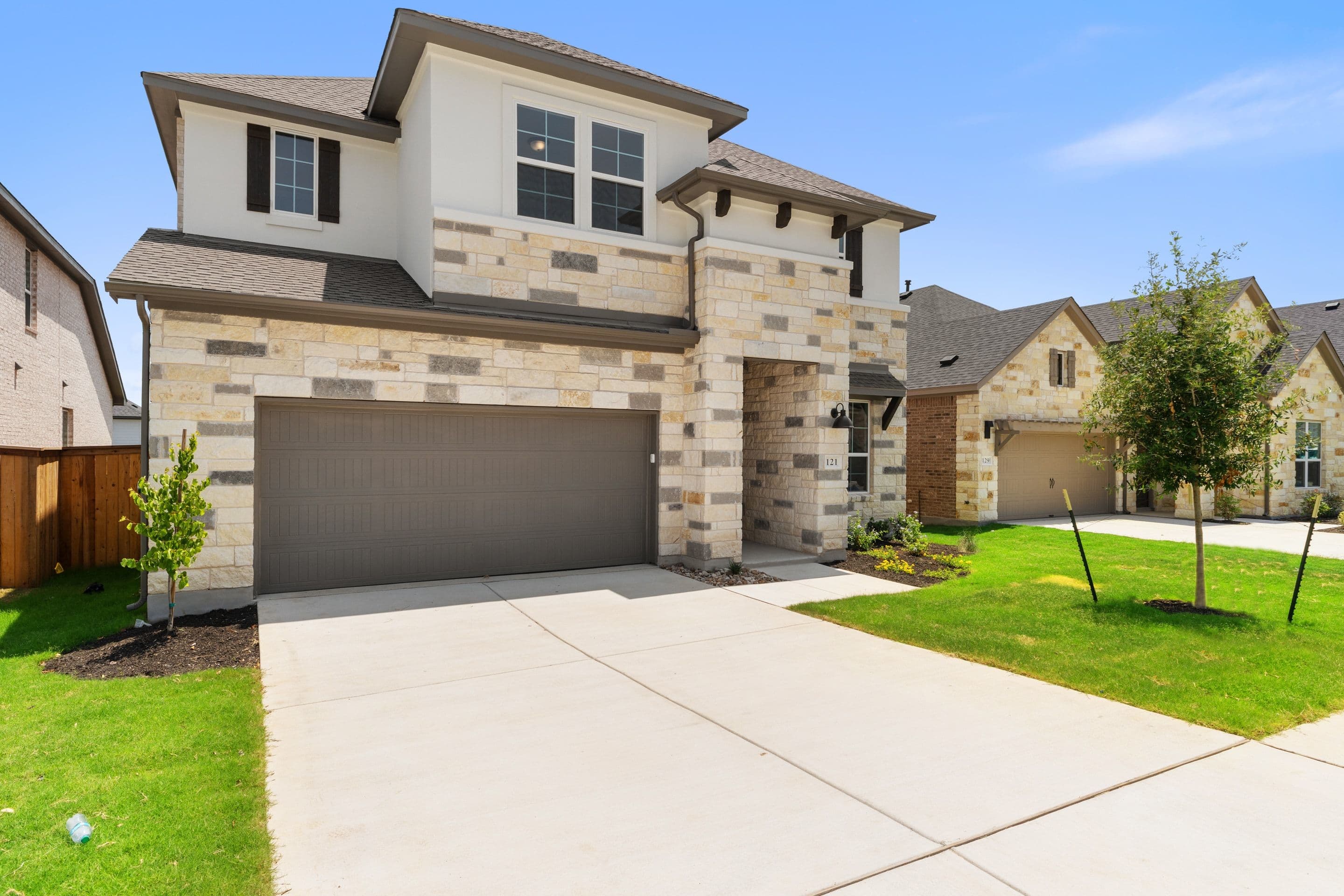 A modern two-story house with stone and stucco exterior and a concrete driveway sits under a clear blue sky, with similar houses lining the street in the background.