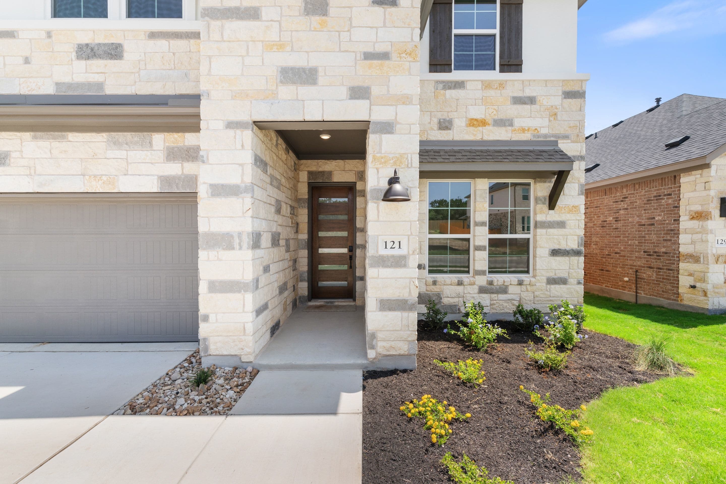 A modern stone-clad house with a glass-paneled door and a manicured lawn sits against a backdrop of similar suburban homes.