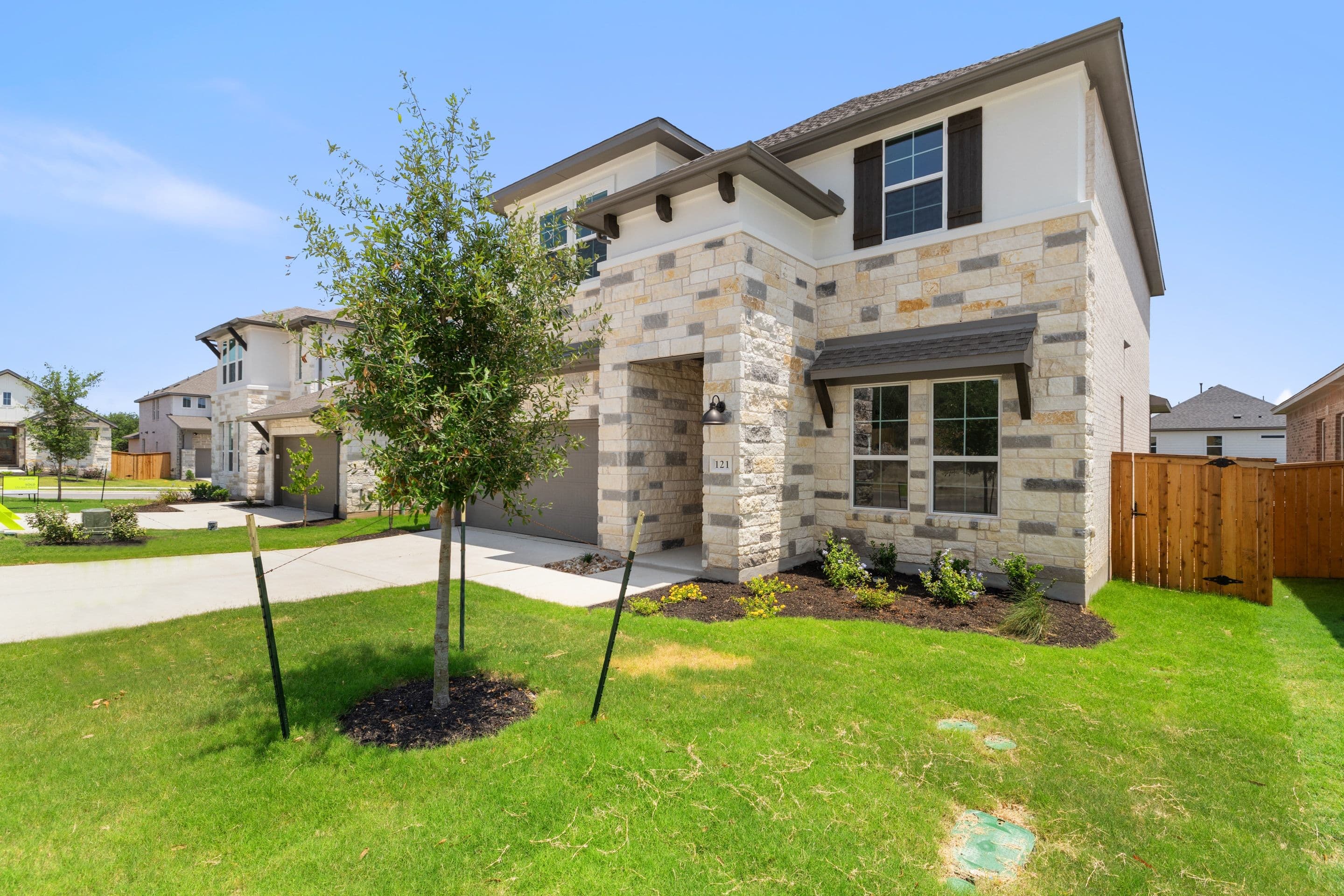 A modern stone-accented house with a manicured lawn in the foreground sits under a clear blue sky, with other similar houses visible in the background.