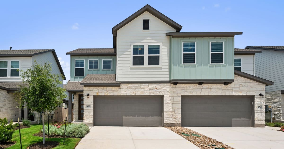 A concrete driveway leads to a row of modern townhouses with stone and light-colored siding, set against a clear blue sky.
