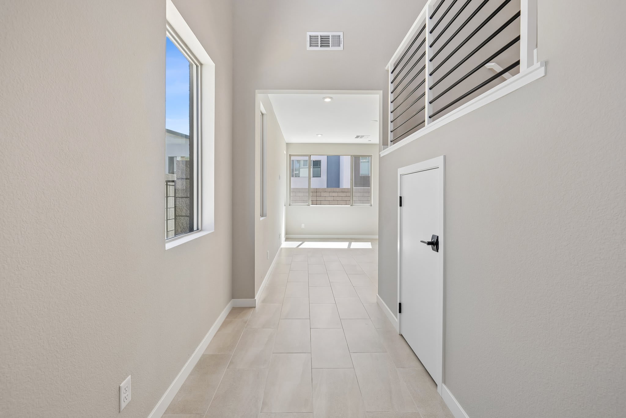 A light-colored hallway with tiled flooring leads to a bright living area with large windows, while a modern staircase with horizontal railings overlooks the space.