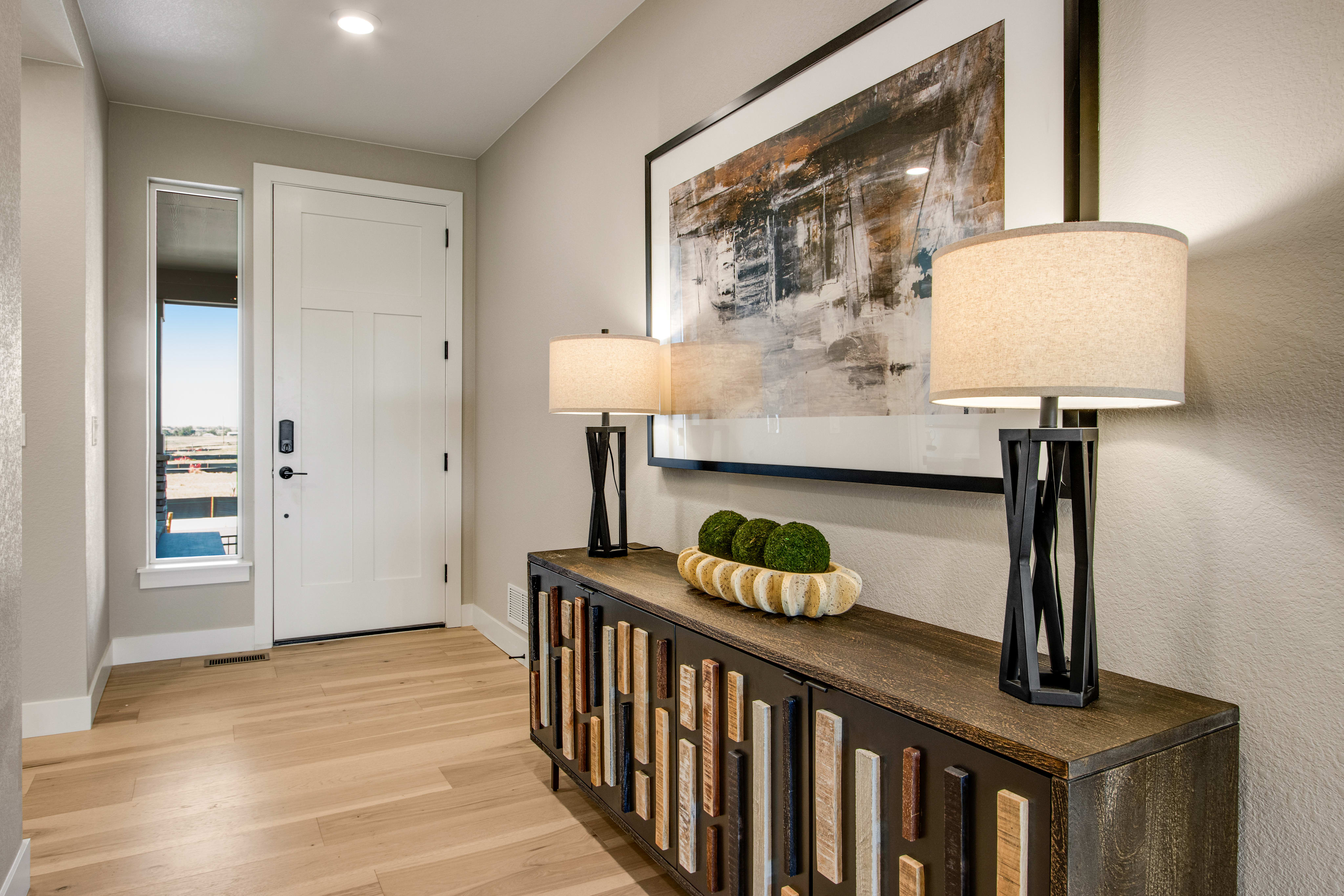 A modern entryway features a wooden console table with decorative lamps and a large abstract painting above it, while a white door and a tall window are visible in the background.