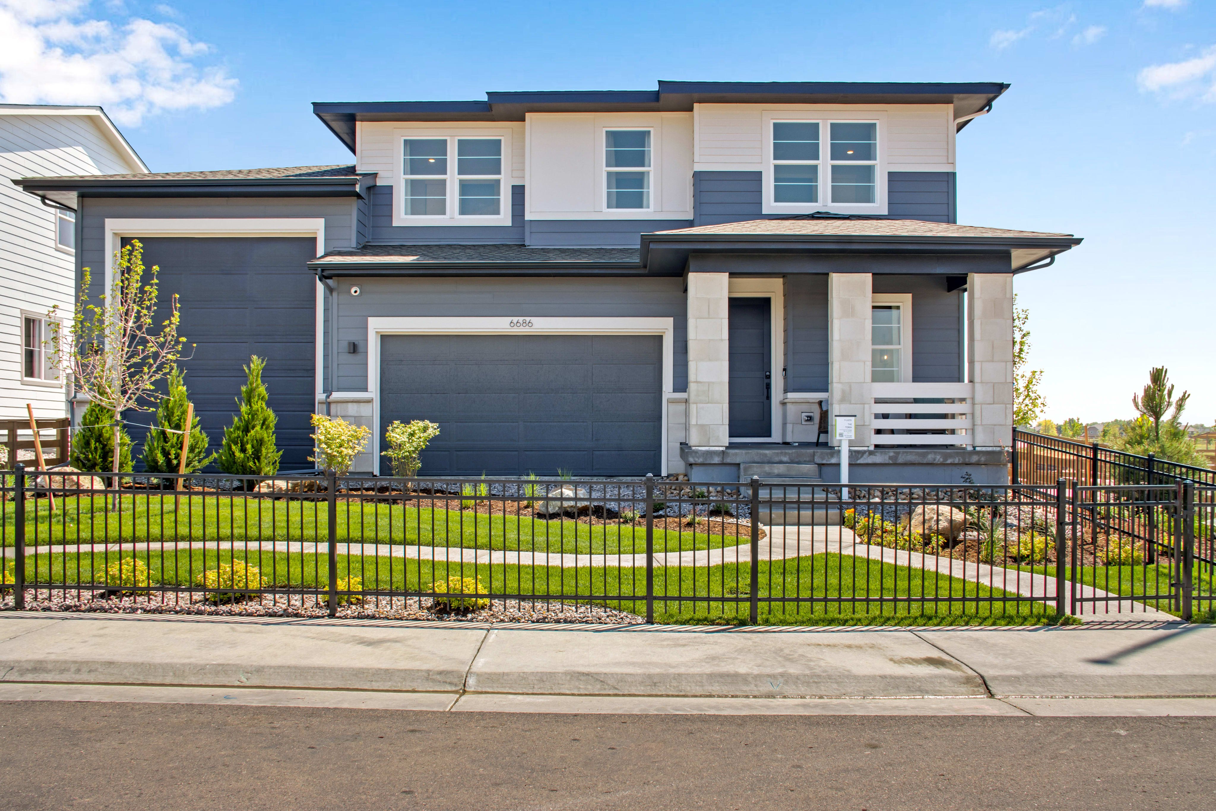 A modern, two-story house with a dark gray and white exterior sits behind a black metal fence and a manicured lawn, with a clear blue sky and distant hills in the background.