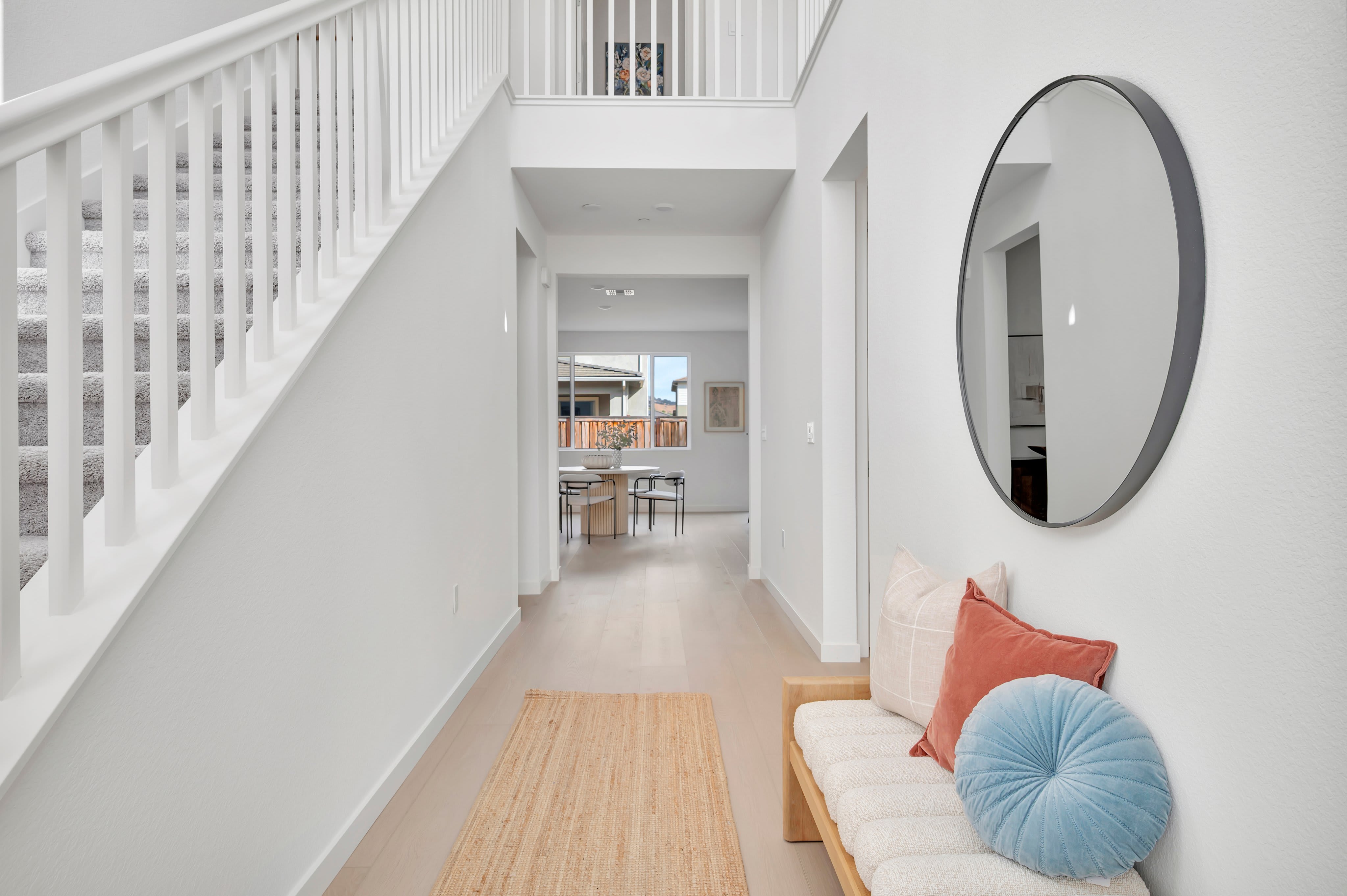 A modern entryway features a white staircase and a wooden bench with colorful pillows in the foreground, leading to a bright dining area visible in the background.